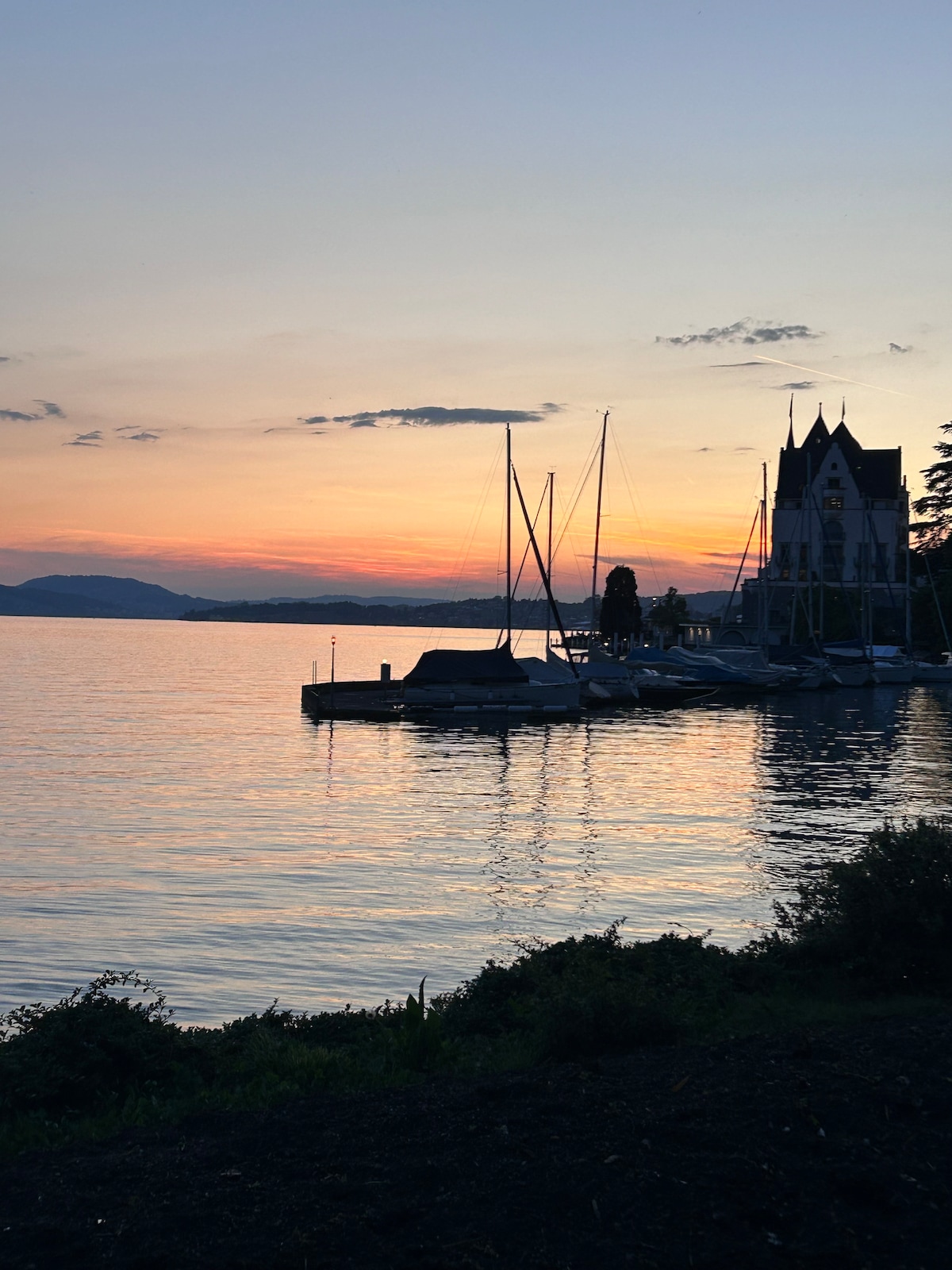 A tranquil lakeside view is captured during sunset, showcasing a calm Lake Lucerne reflecting soft hues of orange and purple. Sailboats are moored at a small dock, with a charming building positioned near the water, surrounded by trees and a scenic backdrop of distant mountains.