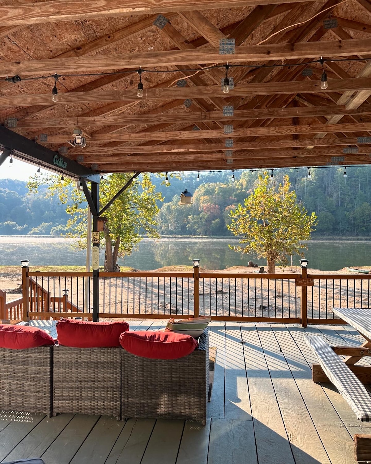 A covered deck features a spacious outdoor seating area with red cushions, overlooking the tranquil New River. Lush greenery can be seen in the background, and the sun casts a warm light on the wooden deck and surrounding area.