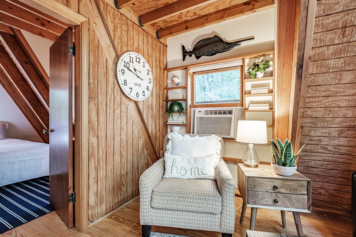 A cozy corner of the cottage features a comfortable armchair with a decorative pillow stating 'home.' A small side table holds a lamp and a potted plant. A large wall clock and shelves are visible, with a glimpse of an air conditioning unit by the window.