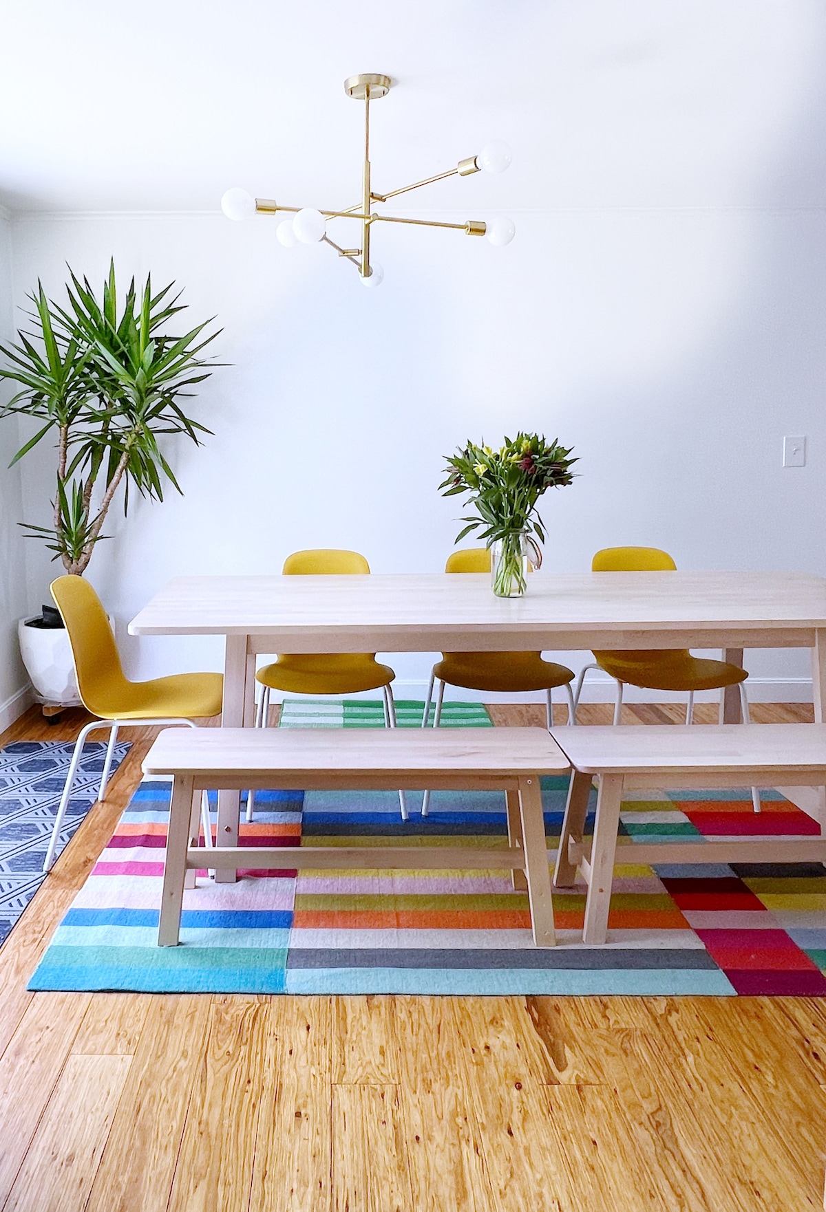 A modern dining area is presented with a light wooden table surrounded by yellow chairs and a long bench. A vibrant, multicolored rug lies underneath, and a bouquet of flowers sits at the center. A tall green plant adds a refreshing touch to the space.