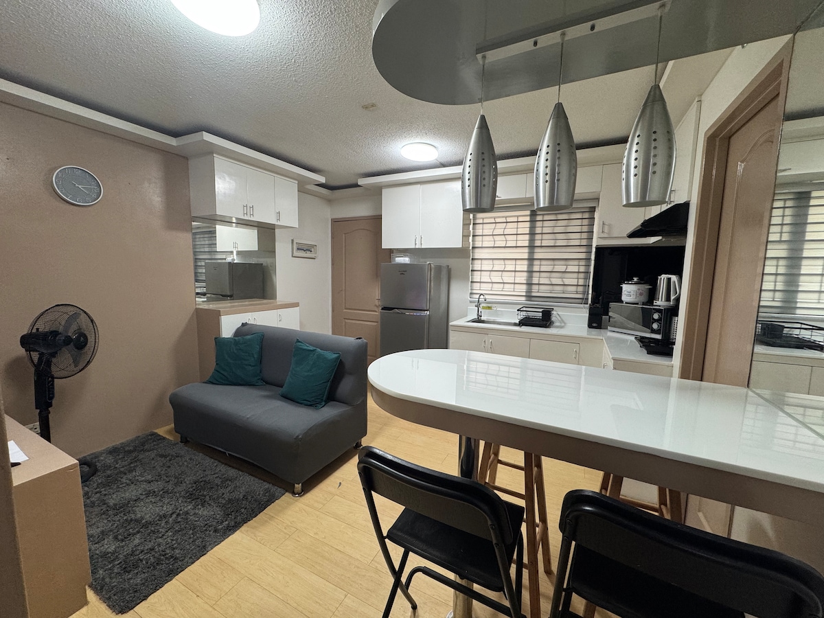 The open living area features a small gray sofa with two teal cushions, accompanied by a round dining table with four black chairs. A compact kitchen area is visible in the background, equipped with a refrigerator and various kitchen essentials. Natural light streams in through the window.