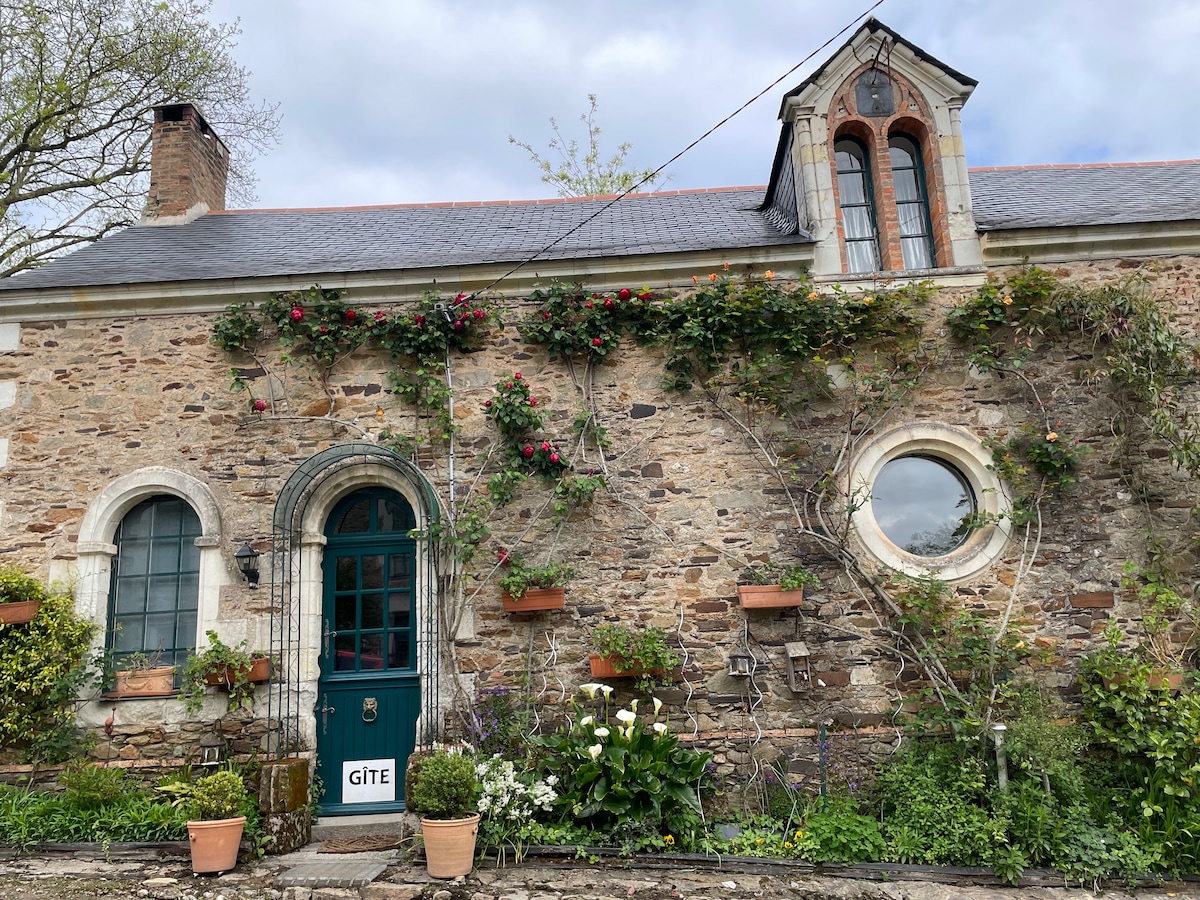 The exterior of a charming stone cottage is highlighted, featuring a green door marked 'GÎTE.' The facade is enhanced by climbing plants and flower pots, with a circular window on one side and a quaint, sloped roof above. Natural greenery surrounds the home.