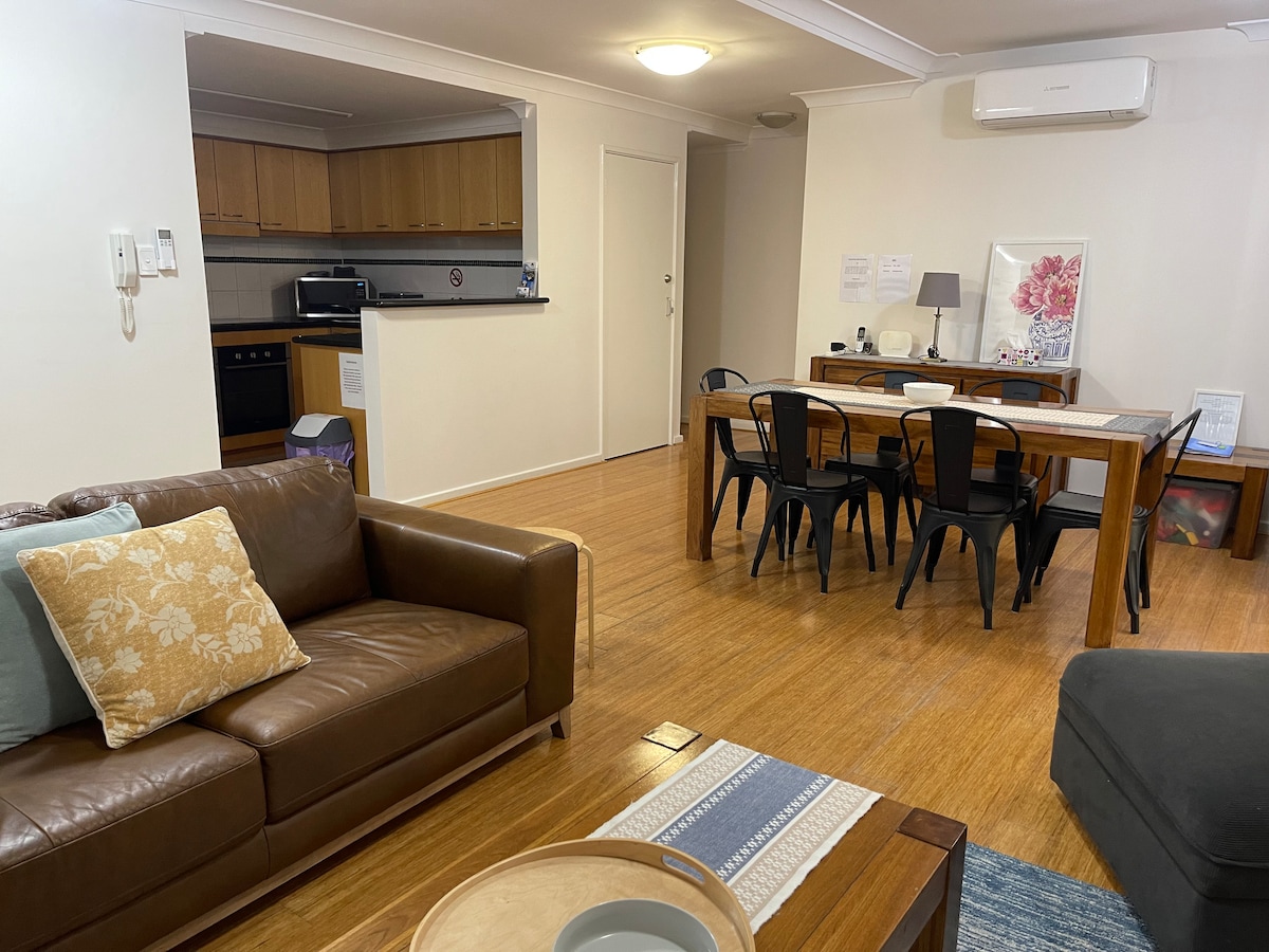 An open-plan living and dining area is showcased, featuring a comfortable brown leather sofa with decorative cushions. A wooden dining table with black chairs sits adjacent to a fully equipped kitchen, while air-conditioning is positioned above to maintain comfort.