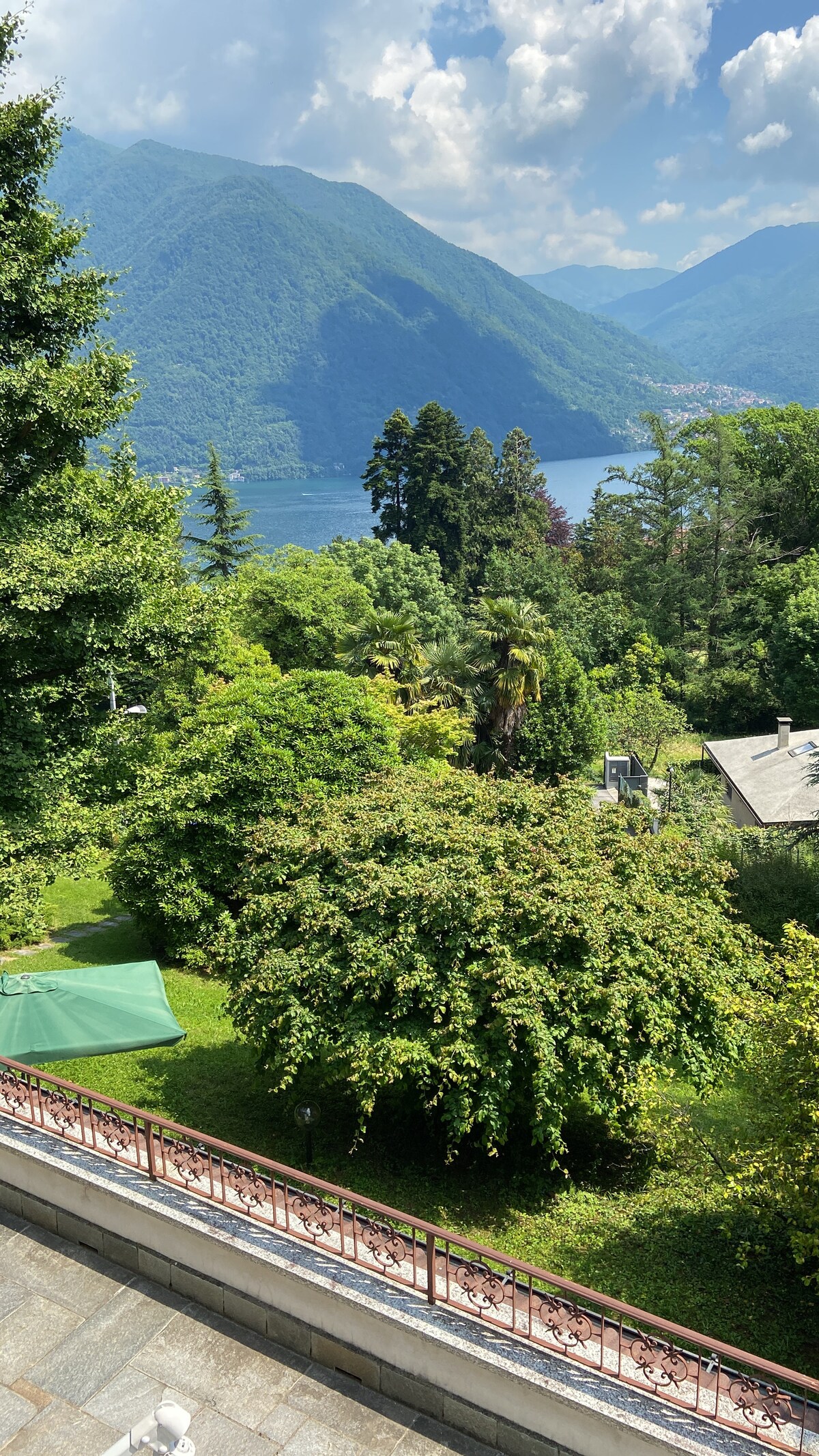 A scenic view captures lush greenery and trees overlooking a serene body of water. The backdrop is formed by impressive mountains under a partially cloudy sky. A tent is visible in the garden area, blending harmoniously with the natural surroundings.