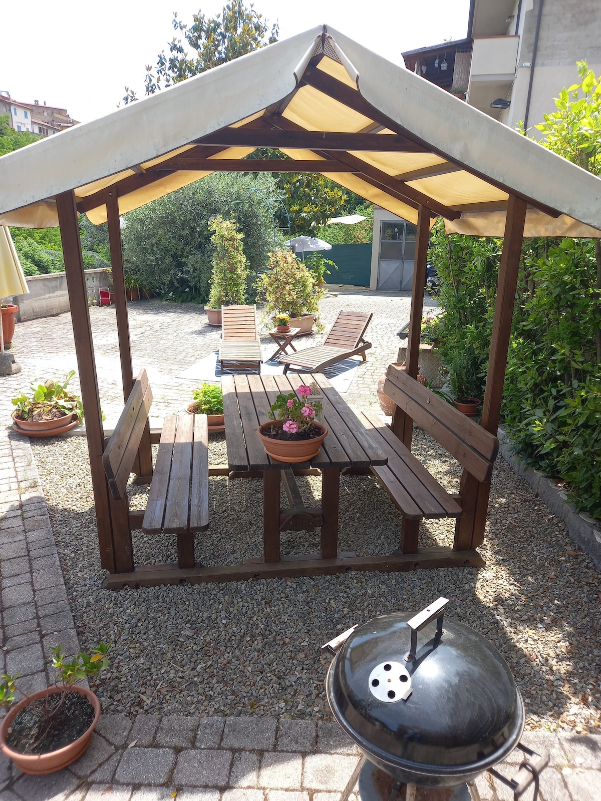 An outdoor shaded seating area is featured with a wooden picnic-style table and benches. Potted flowers add color, while a grill is positioned nearby. The area is surrounded by greenery and stone pathways, creating an inviting space for outdoor dining and relaxation.