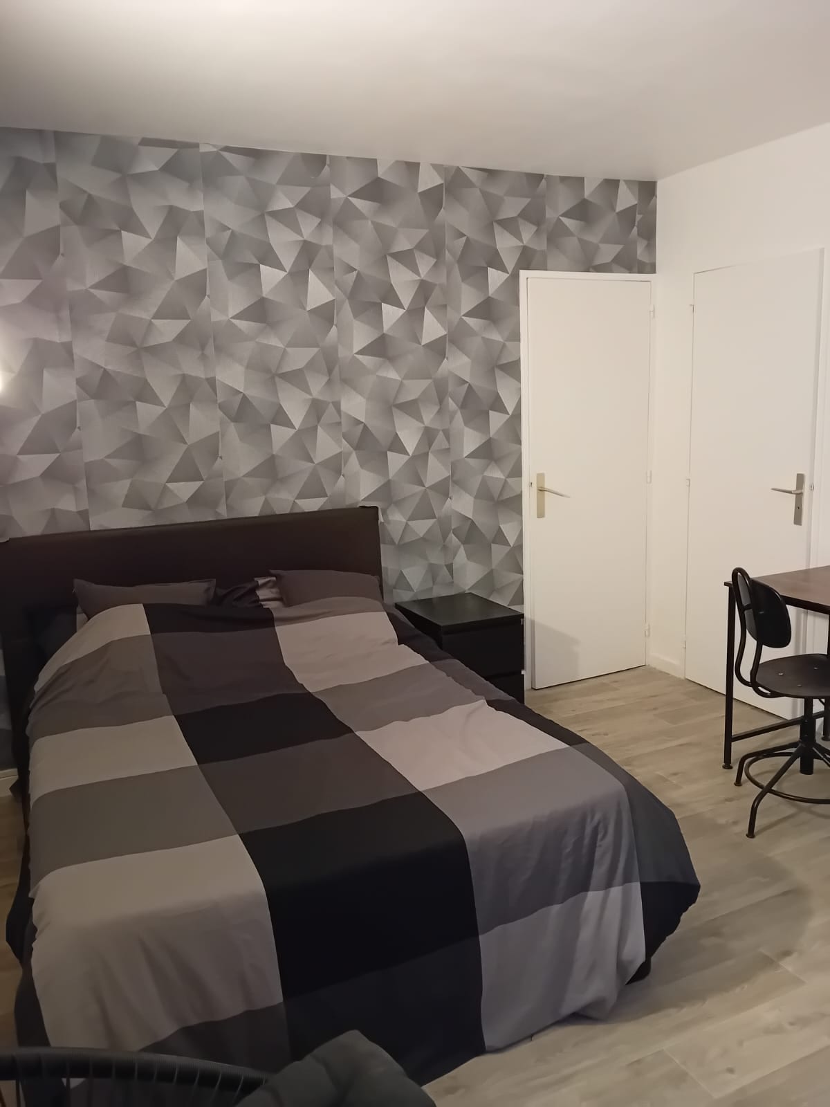 A modern sleeping area features a spacious bed dressed in a geometric patterned duvet. Adjacent, a small black nightstand complements the decor, and a minimalist desk with a chair is positioned nearby. The walls are decorated with textured wallpaper in shades of gray.
