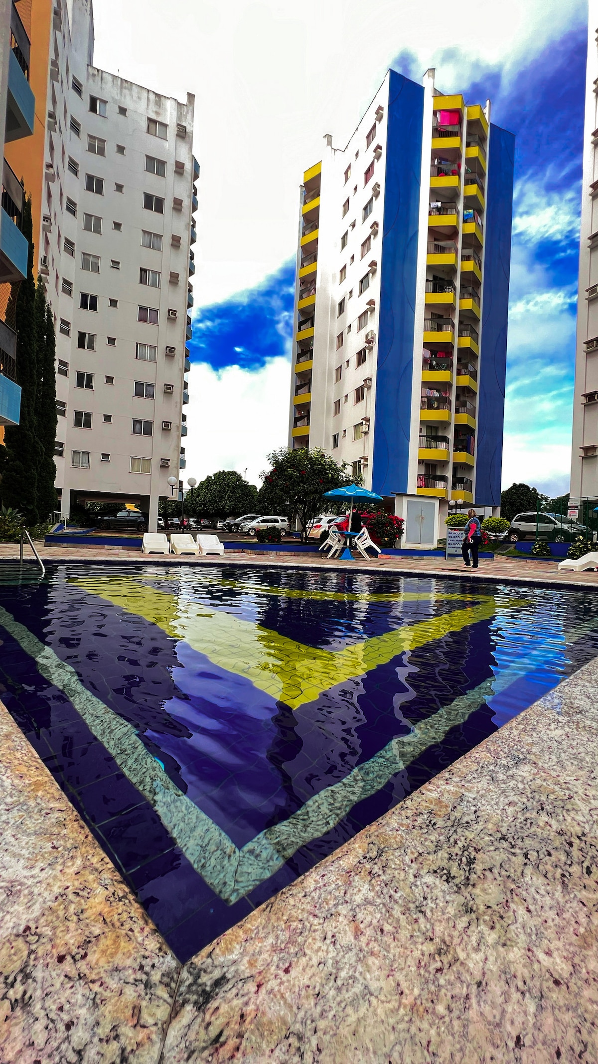 A spacious outdoor pool is framed by colorful buildings that rise in the background. Lounge chairs are arranged alongside the water, reflecting the vibrant hues from the surrounding architecture. Clear skies are visible overhead, enhancing the inviting atmosphere of the area.
