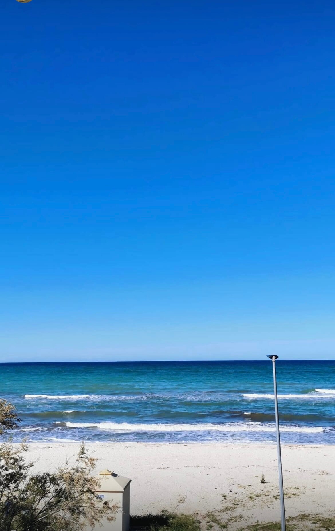 A clear blue sky displays a gentle gradient down to the sea, where smooth waves roll onto a sandy beach. The image captures the serene water in various shades of turquoise, enhancing the overall calm ambiance of the coastal scene.
