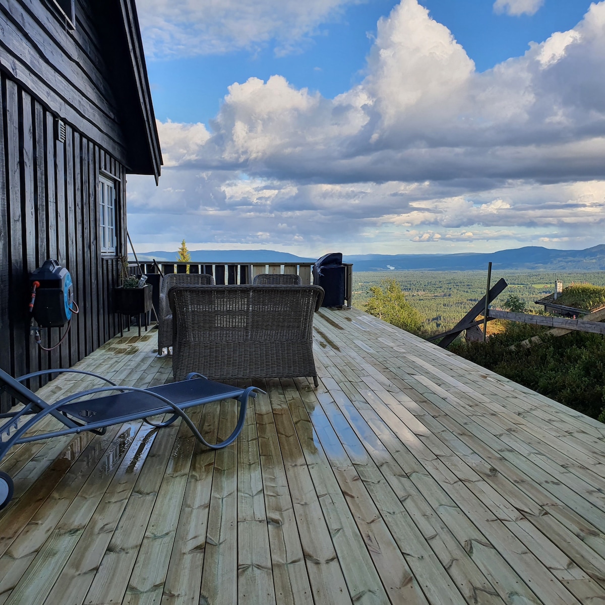 A spacious wooden terrace extends from the property, offering expansive views of the surrounding mountains and valleys. Comfortable outdoor seating is visible, with a chaise lounge positioned for relaxation. The terrace surface appears clean and well-maintained, reflecting the sky's light after recent precipitation.