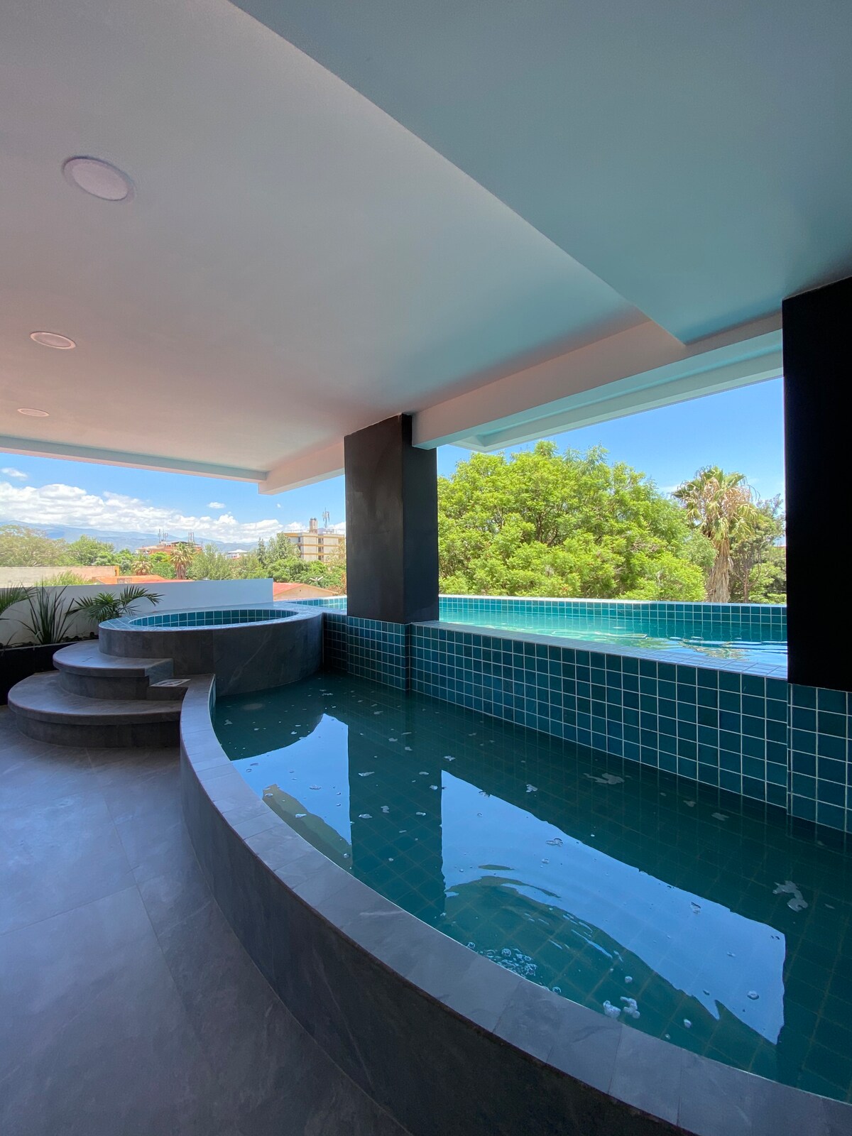 An inviting shared pool area features a curved design with steps leading into the shallow water. Large windows offer views of the surrounding greenery and blue sky, creating a serene atmosphere. The pool is surrounded by modern tile accents and is situated in a well-lit space.