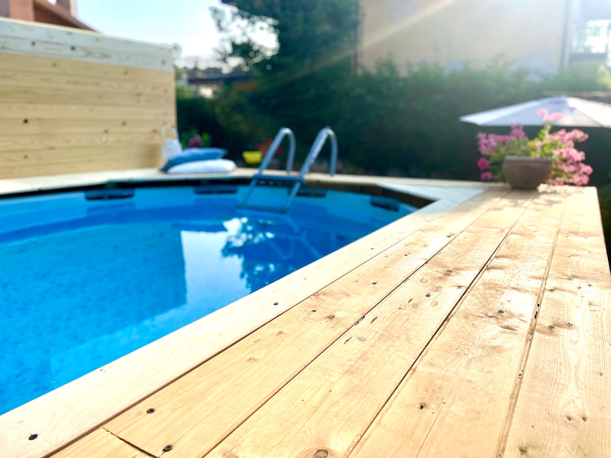 The image features a circular above-ground pool surrounded by a wooden deck. Sunlight reflects off the clear blue water, and flowering plants provide a touch of color. A ladder leads into the pool, while a sun umbrella stands nearby, creating a relaxing outdoor atmosphere.