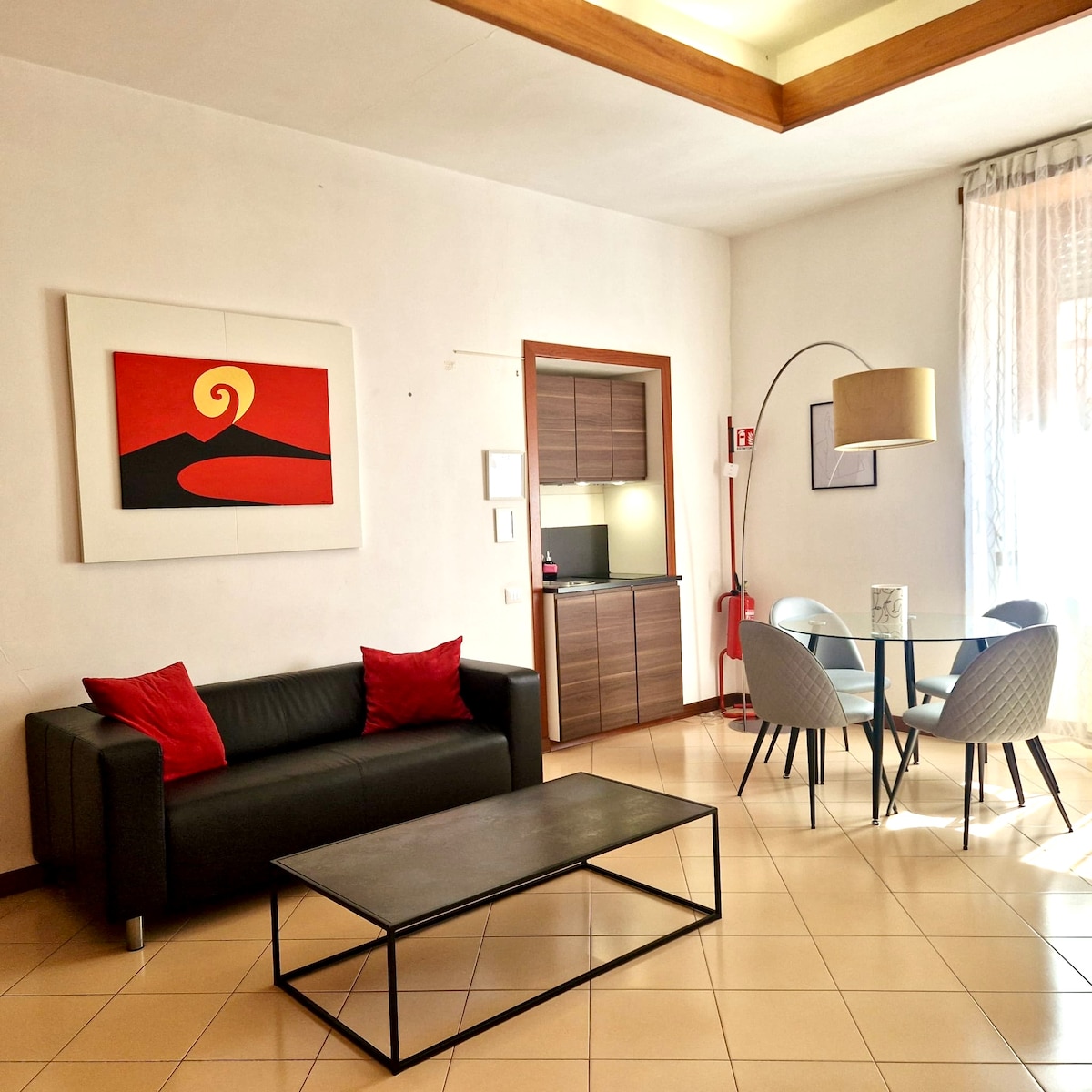 A contemporary living space is showcased, featuring a dark couch adorned with red cushions. A glass table with grey chairs is positioned in front of a large window, which allows natural light to brighten the room. A painting adds a touch of color to the wall, complementing the functional kitchenette in the background.