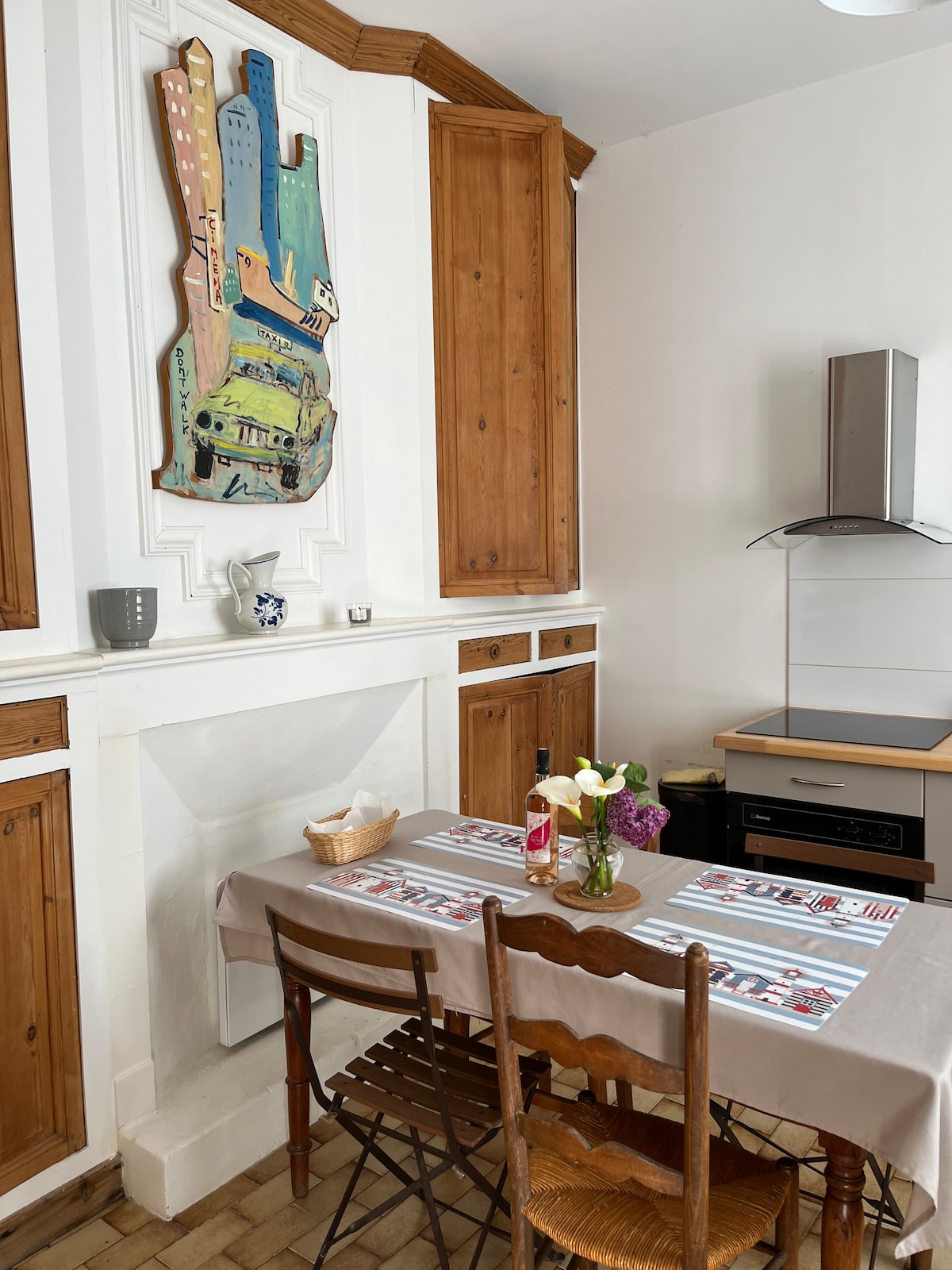 The kitchen area displays a small wooden dining table set for four, complemented by simple chairs. A colorful wall art piece adds visual interest above a well-kept fireplace. Wooden cabinetry provides storage, and a modern stovetop is positioned nearby, contributing to a functional cooking space.