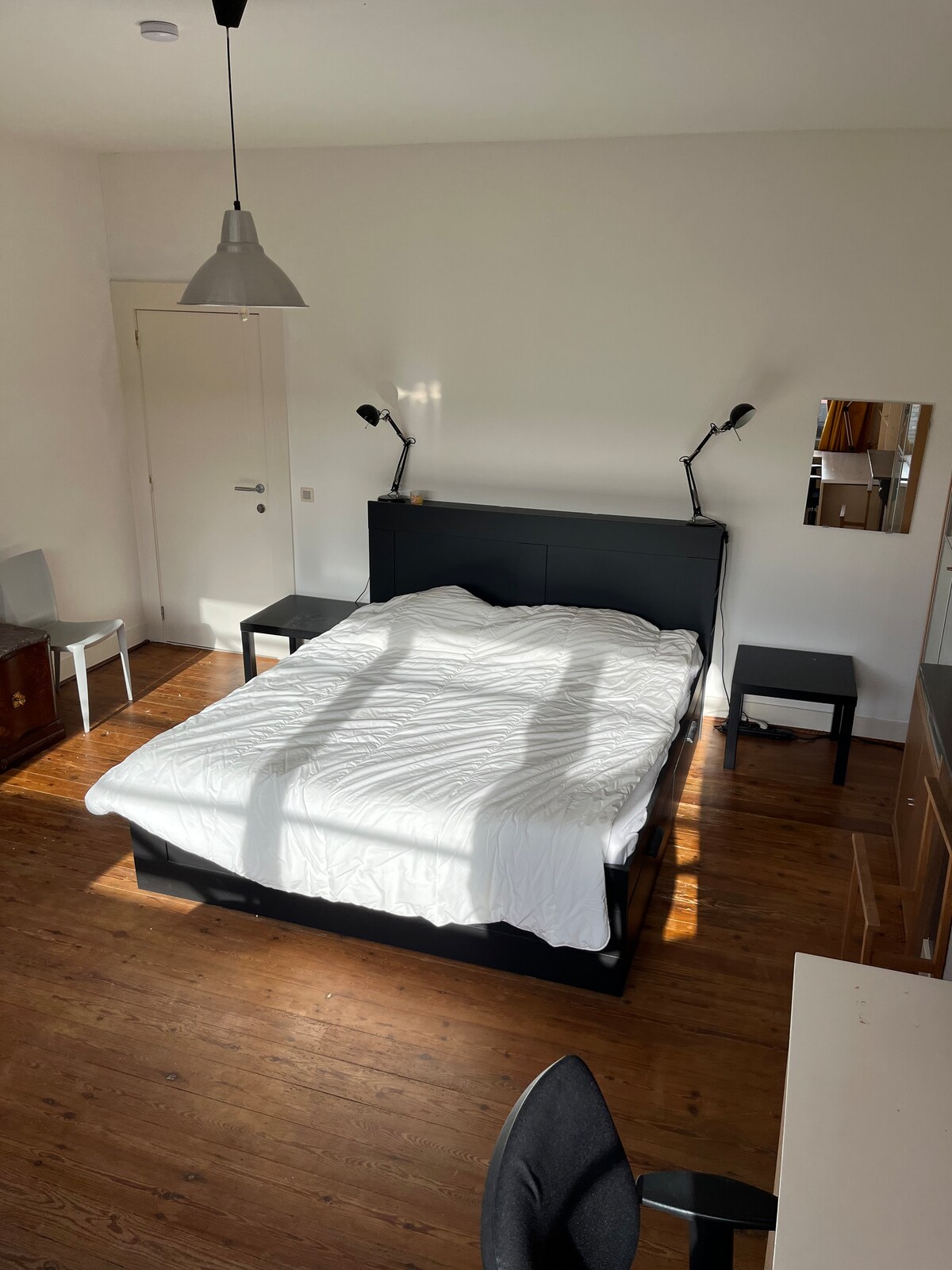 A spacious bedroom features a large bed with a crumpled white comforter. Two bedside lamps flank the bed, providing task lighting. A wooden floor reflects natural light streaming through the window, complemented by a simple desk and a dresser against the wall.