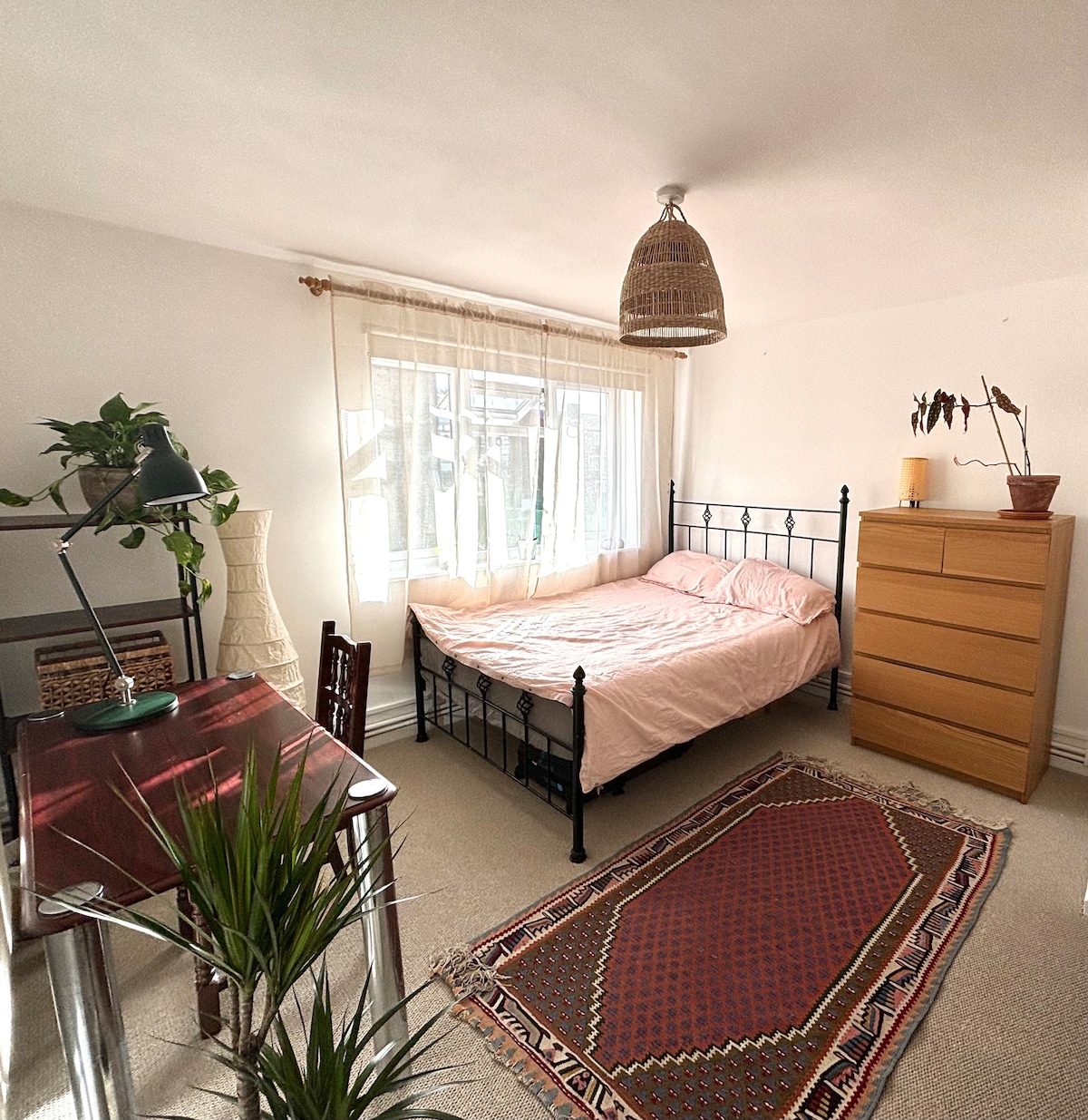 A spacious bedroom features a metal frame bed with soft pink bedding. Natural light floods the room through a large window, complemented by a wooden dresser and a small desk with a desk lamp. Indoor plants add a touch of greenery, and a patterned rug enhances the floor.