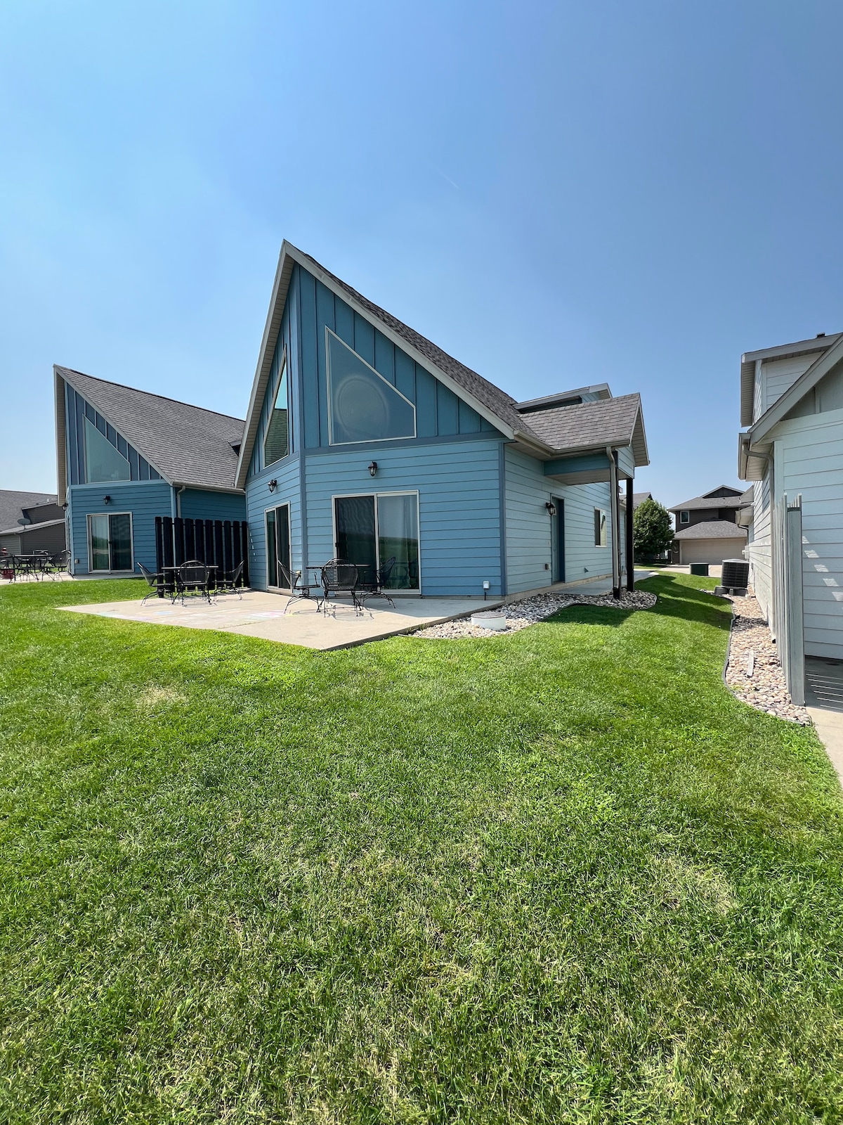A vacation cabin with a light blue exterior is shown, featuring a triangular roof and large windows. The grassy yard surrounds a patio area with several outdoor chairs. Adjacent cabins are visible, creating a welcoming atmosphere for larger groups.