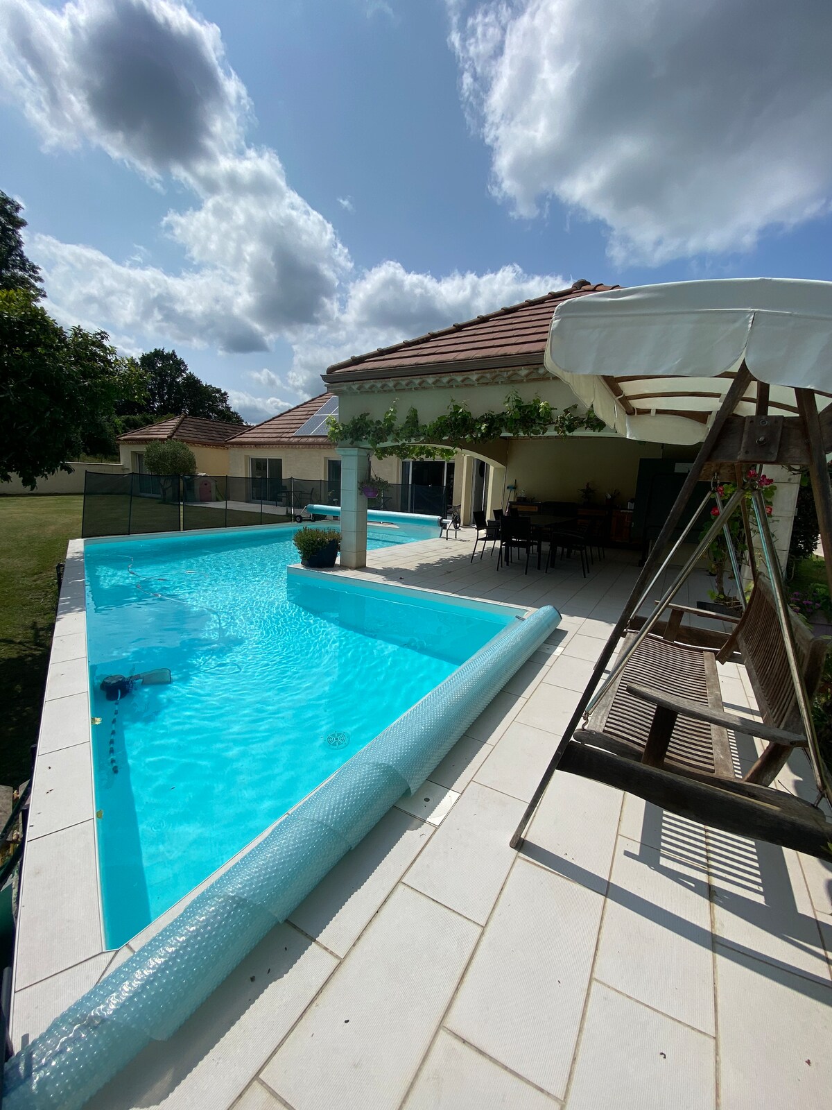 A private swimming pool, surrounded by a tiled patio, reflects the bright blue sky. A shaded area with a swing is visible, along with dining furniture under a canopy. Lush greenery frames the space, complementing the tranquil atmosphere of the villa.