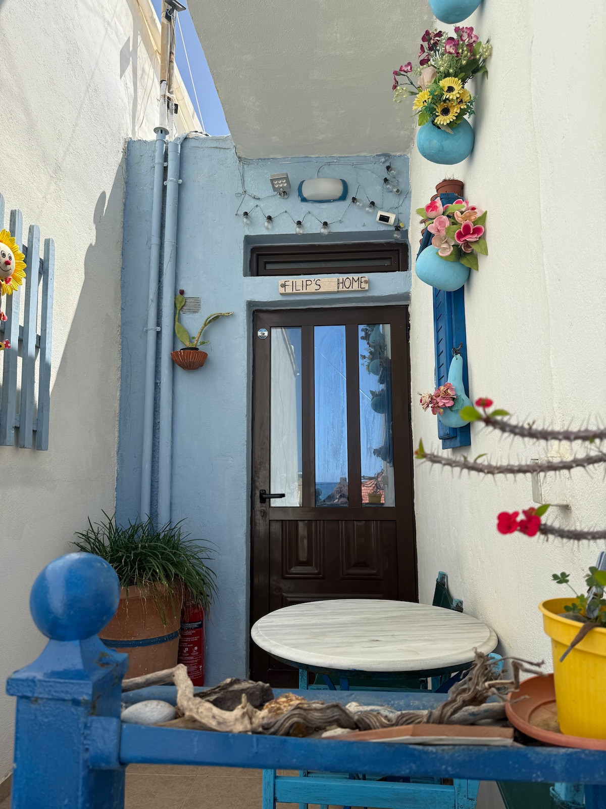 A charming private entrance is framed by light blue walls adorned with colorful flower pots. The wooden door, labeled 'Philips Home,' features a small window above it. A round table with a marble top is situated nearby, complemented by vibrant potted plants and decorative elements.