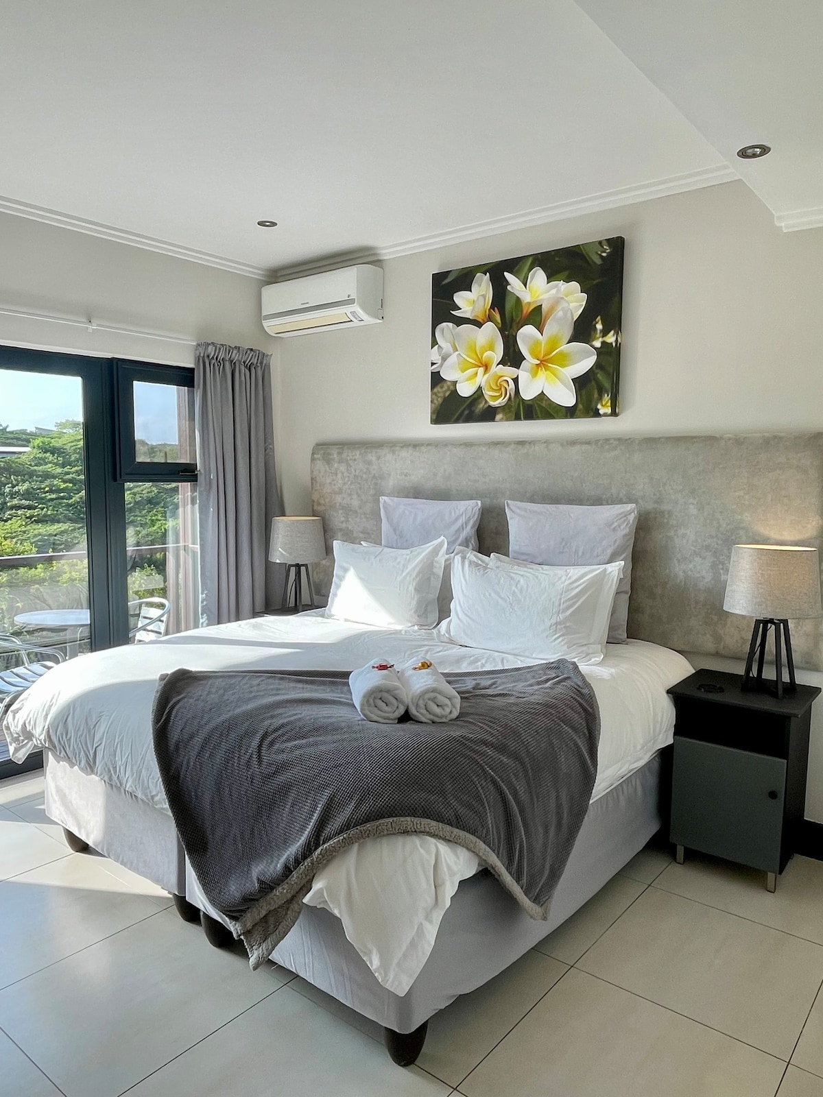 A well-lit bedroom features a spacious bed with white linens and decorative pillows. Two bedside lamps stand on either side. A piece of floral art adorns the wall above the bed. Large windows allow natural light and offer views of the surrounding greenery.