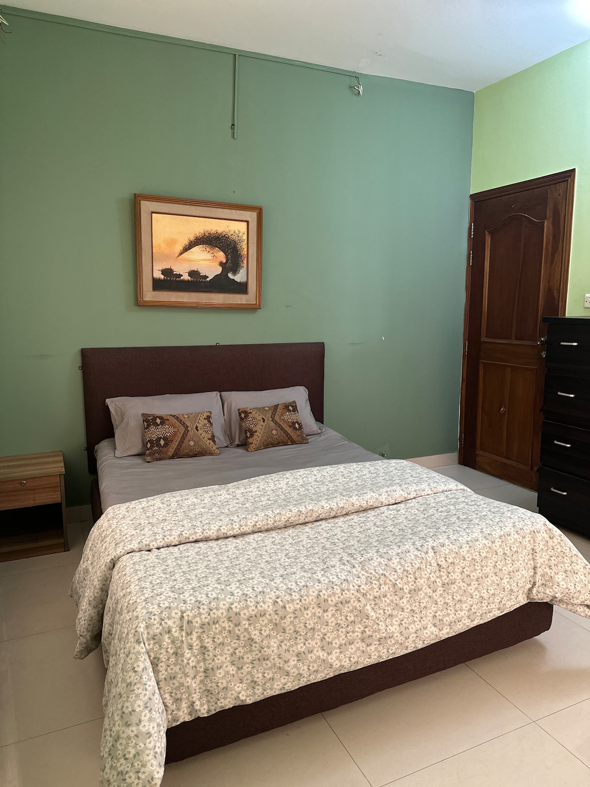 A spacious bedroom is presented with a comfortable bed dressed in gray linens and a patterned quilt. Decorative pillows add a touch of style. A framed artwork adorns the green wall, while a wooden nightstand and dresser offer essential storage.