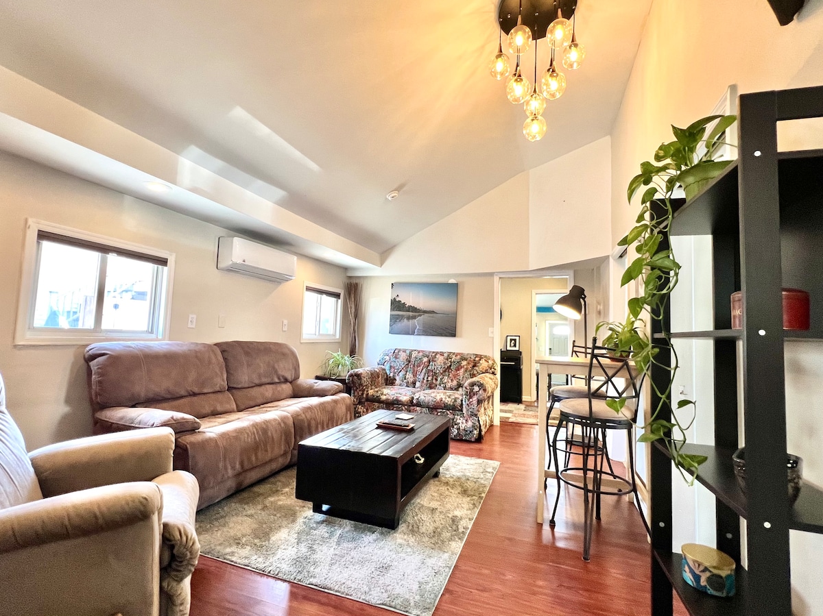 The living area features vaulted ceilings and a mix of comfortable seating, including a large sofa and loveseat. A coffee table sits at the center, complementing the decorative rug. Natural light enters through windows, and a vibrant wall art adds color to the space.
