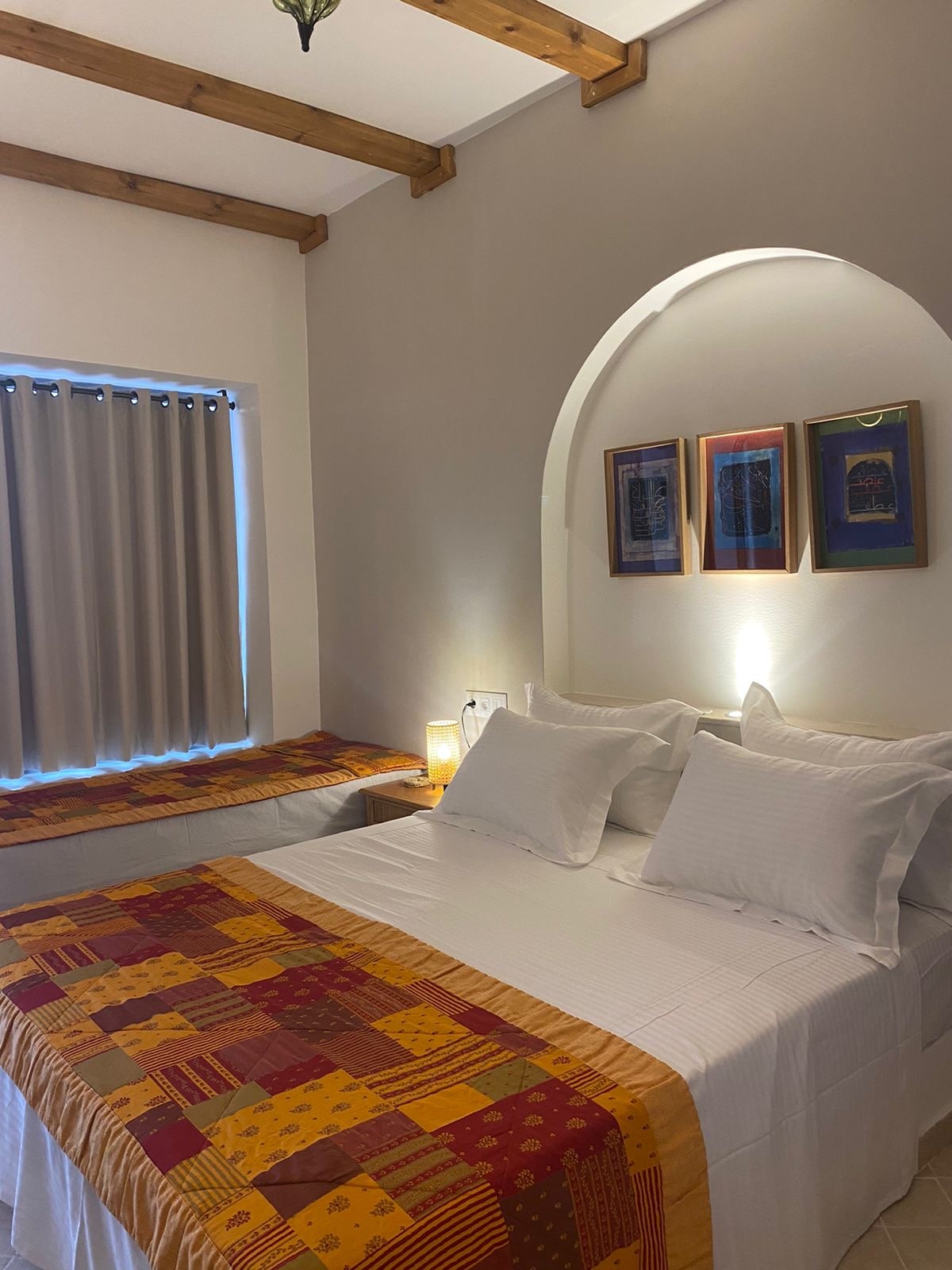 A cozy room is presented with a spacious bed and an additional single bed. Colorful patterned bedspreads add a warm touch. Soft light is emitted from a bedside lamp, and framed artwork adorns the wall, enhancing the inviting atmosphere of the space.