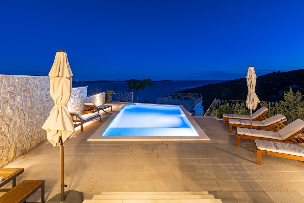 A serene outdoor area features a well-lit swimming pool surrounded by sun loungers. Soft umbrellas stand beside the loungers, ensuring comfort during sunny days. The tranquil sea view is visible in the background, with gentle evening light enhancing the peaceful atmosphere.
