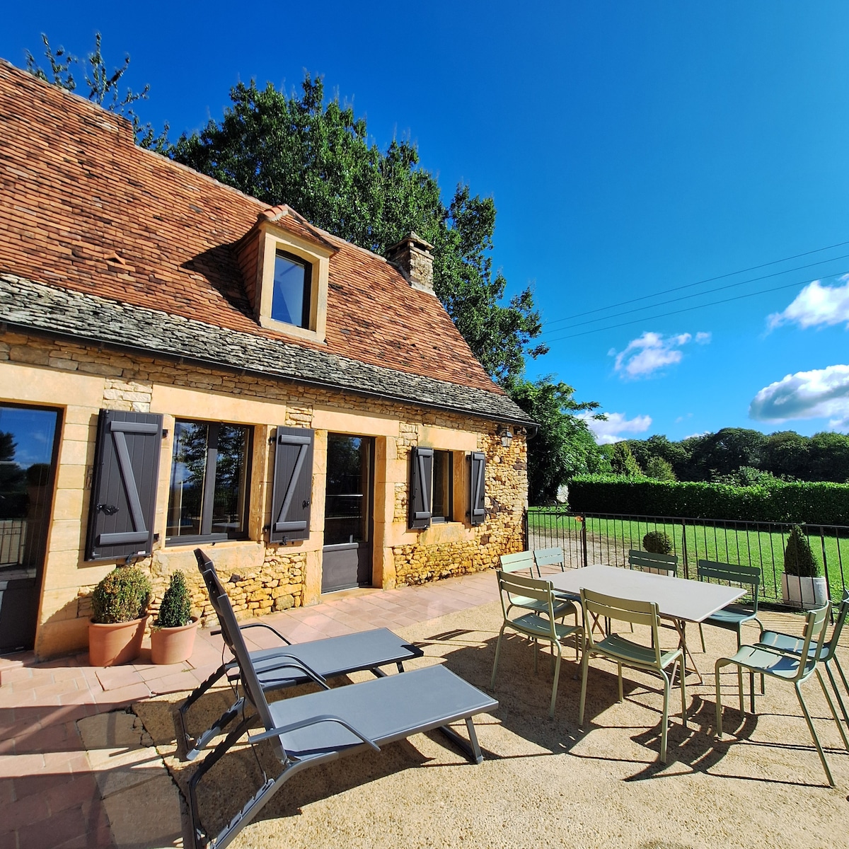 The exterior view features a stone house with a distinctive roof and landscaped surroundings. A seating area includes a table and chairs, complemented by lounge chairs. Green hedges and a clear blue sky enhance the tranquil environment, creating a welcoming outdoor space.