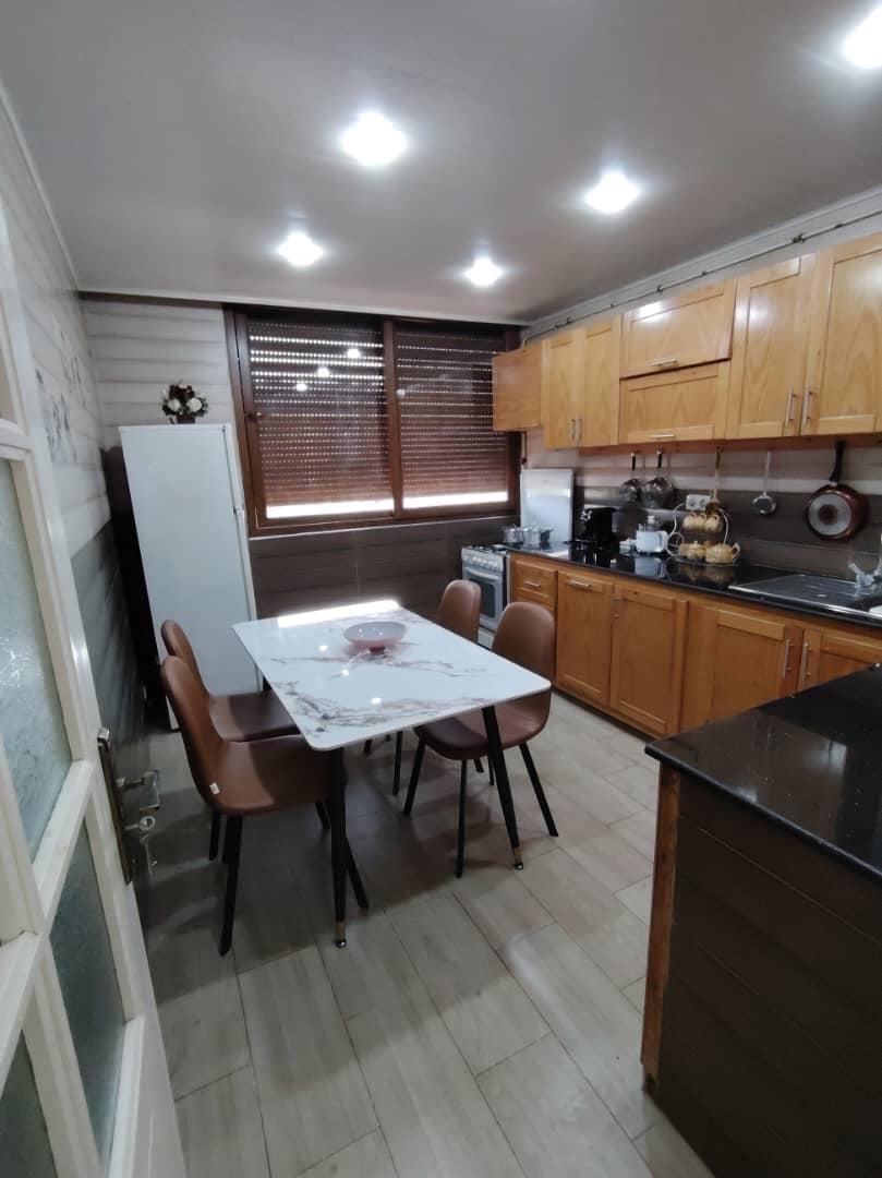 A modern kitchen features wooden cabinets and a central table with a marble top surrounded by four chairs. Large windows with blinds allow natural light to enter, and kitchen appliances are neatly arranged along the walls, creating an organized and functional space.