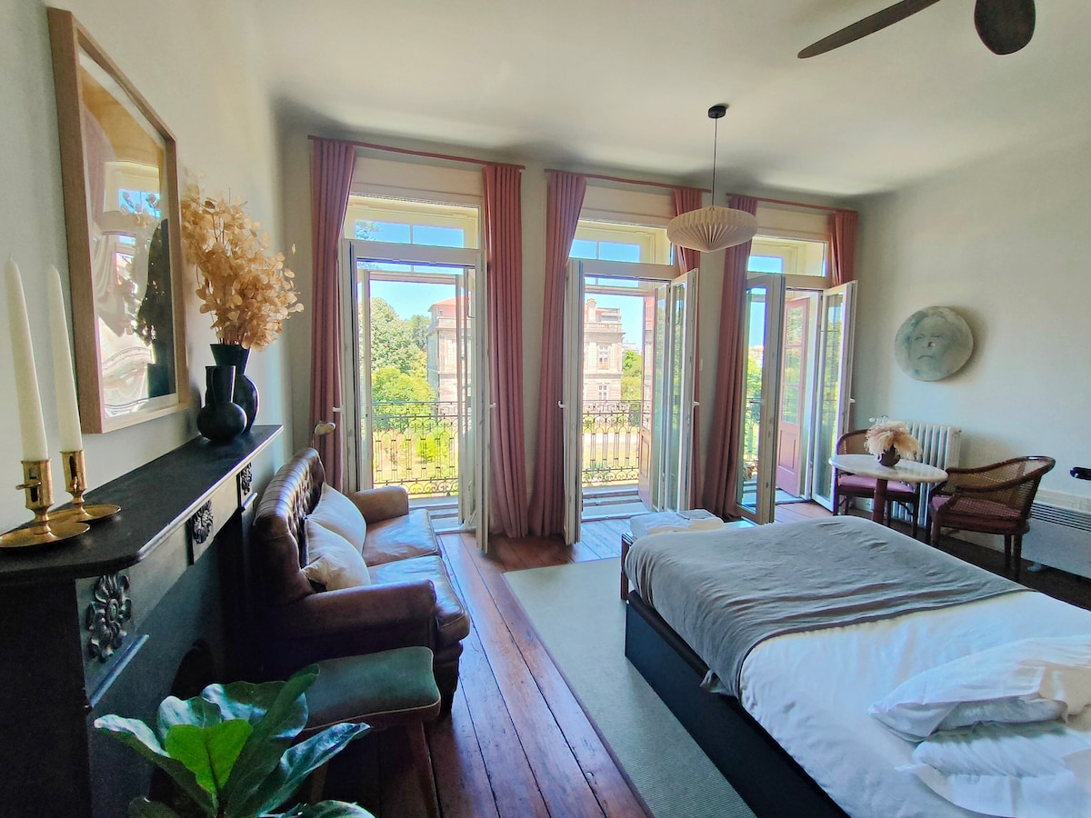 A spacious suite is displayed, featuring a comfortable bed with a neutral-toned coverlet. Large windows with soft drapes provide natural light, overlooking a scenic view. A seating area with a small table and a decorative wall piece add character to the inviting space.