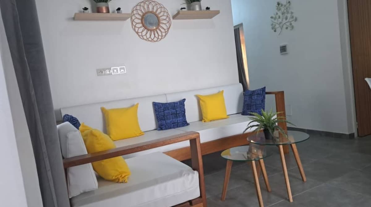 A bright living area is furnished with a wooden couch featuring multiple yellow and blue cushions. A glass coffee table sits in the center, complemented by a small green plant. Light enters through a nearby doorway, creating a welcoming ambiance.