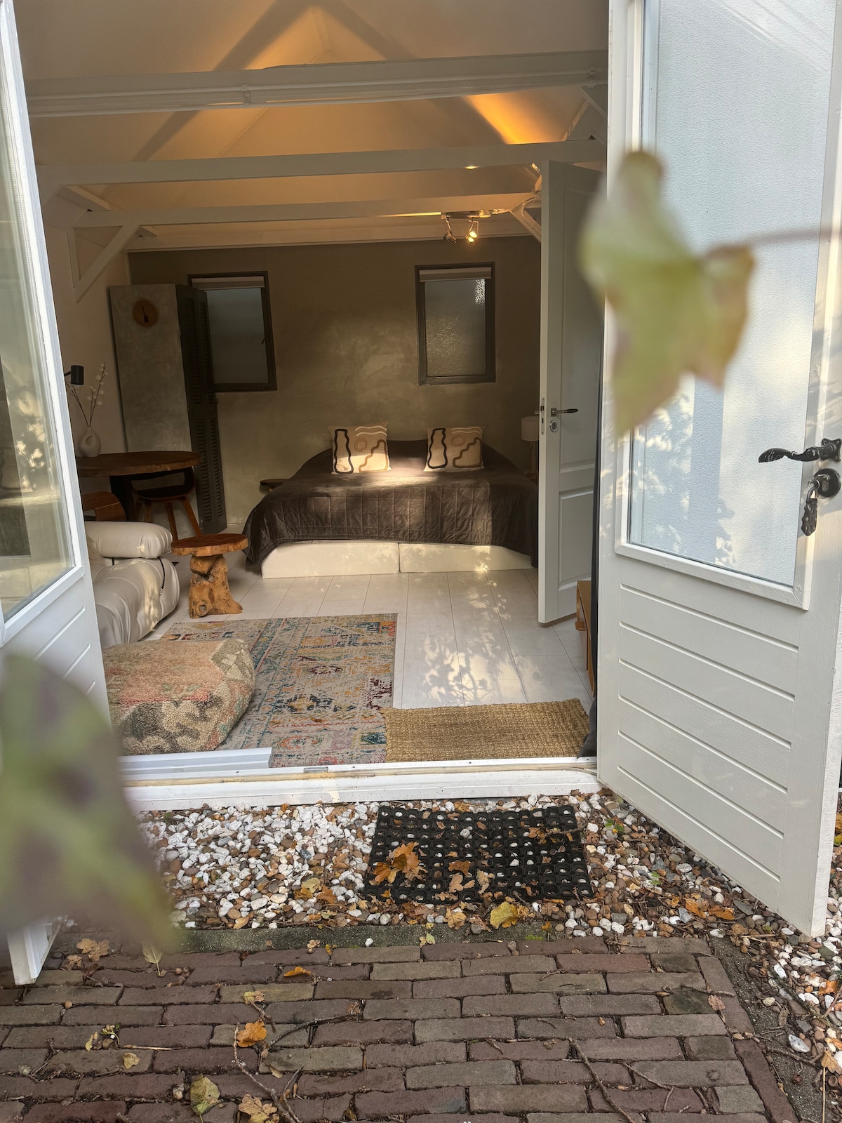 A welcoming entrance opens into a bright sleeping area featuring a spacious bed with soft linens. Natural light fills the room, highlighting the minimalist decor and a subtle color palette. A comfortable seating area is visible, complemented by a decorative rug.