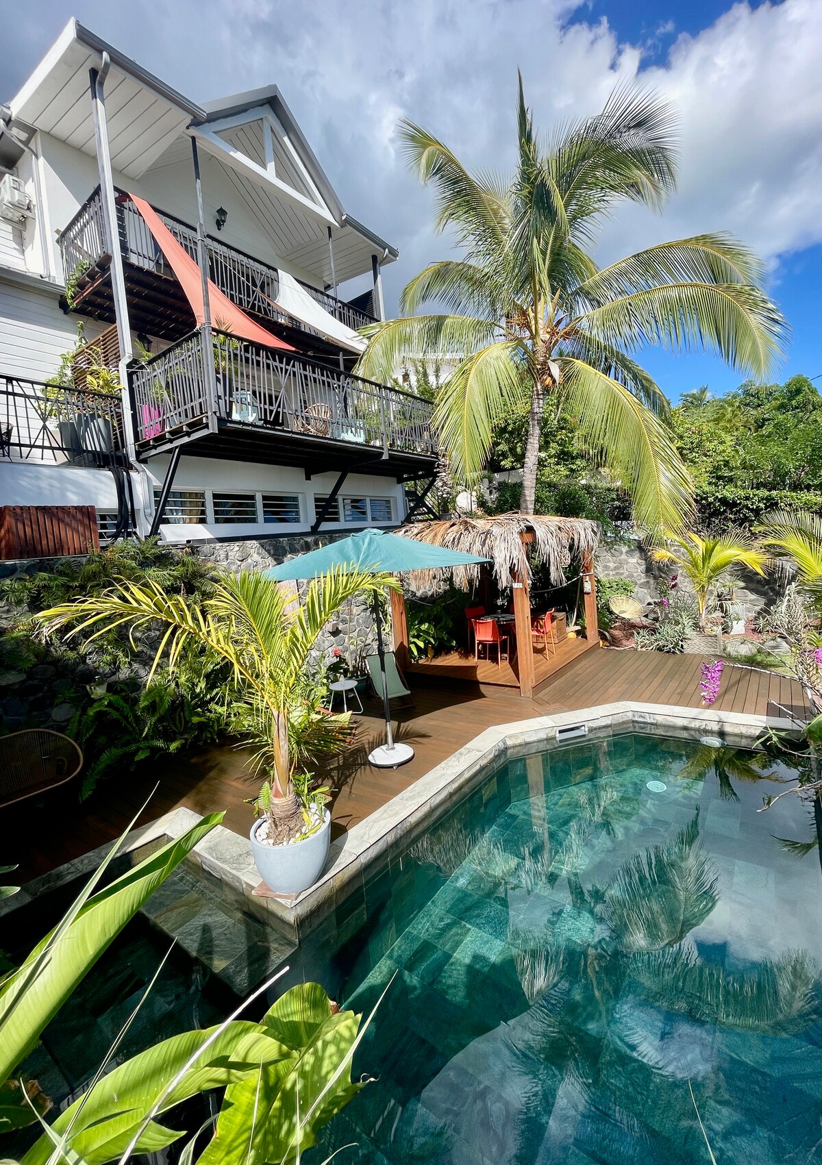 A private pool area is surrounded by tropical plants, with a thatched-roof gazebo providing shade. The water is crystal clear, reflecting the blue sky. A dining area is visible under the gazebo, inviting relaxation amidst the serene outdoor setting.