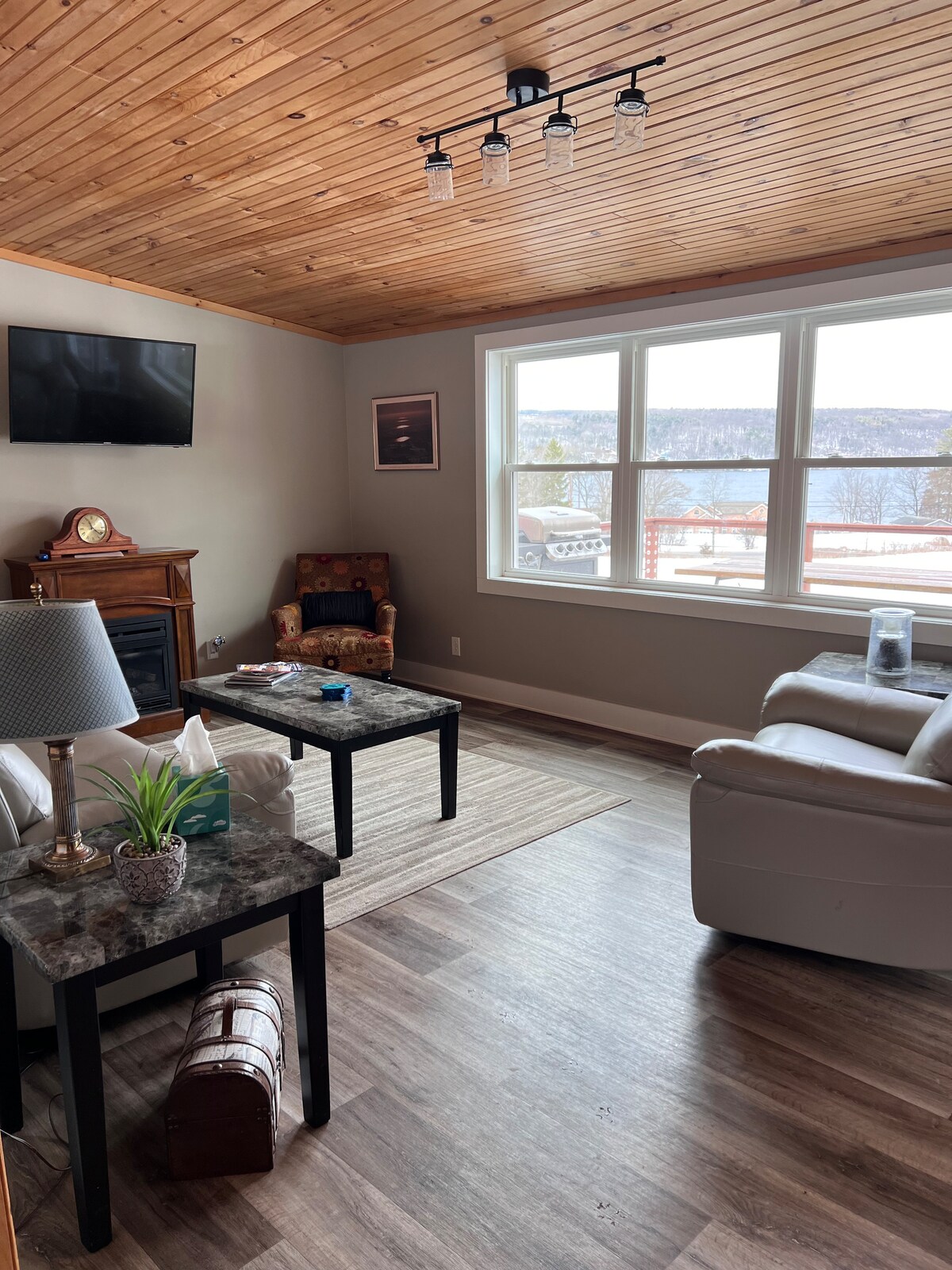 A comfortable living area is highlighted by a wooden ceiling and large windows that allow natural light. A flat-screen television is mounted on the wall, while a small side table with a lamp and a decorative plant complements the seating arrangement, featuring a cozy armchair and a leather sofa.