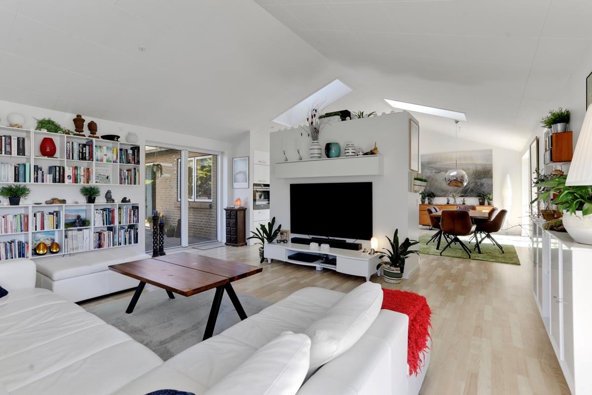 The spacious living area features a white sectional sofa and a wooden coffee table, framed by built-in shelves filled with books. A large TV stands on a sleek white media console. Natural light floods through skylights and glass doors leading to an outdoor space.