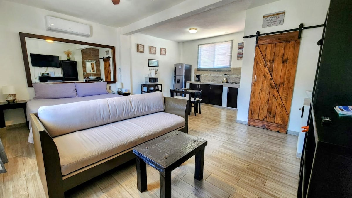 The studio features a spacious layout with a comfortable sofa bed facing a wooden coffee table. A king-size bed is positioned against a large mirror, while a kitchenette and dining area are visible in the background. Natural light fills the space through a nearby window.