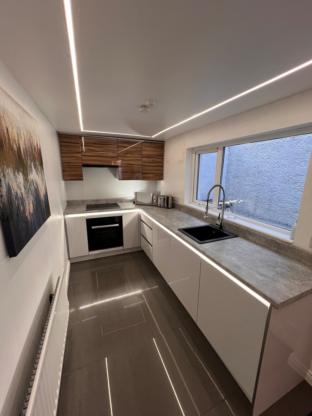 The kitchen is equipped with modern appliances, including an oven and microwave. Cabinets in a warm wood tone contrast against the sleek white cabinetry. A dark sink is positioned under a window, with ample counter space illuminated by linear lighting above and reflecting off the polished floor.