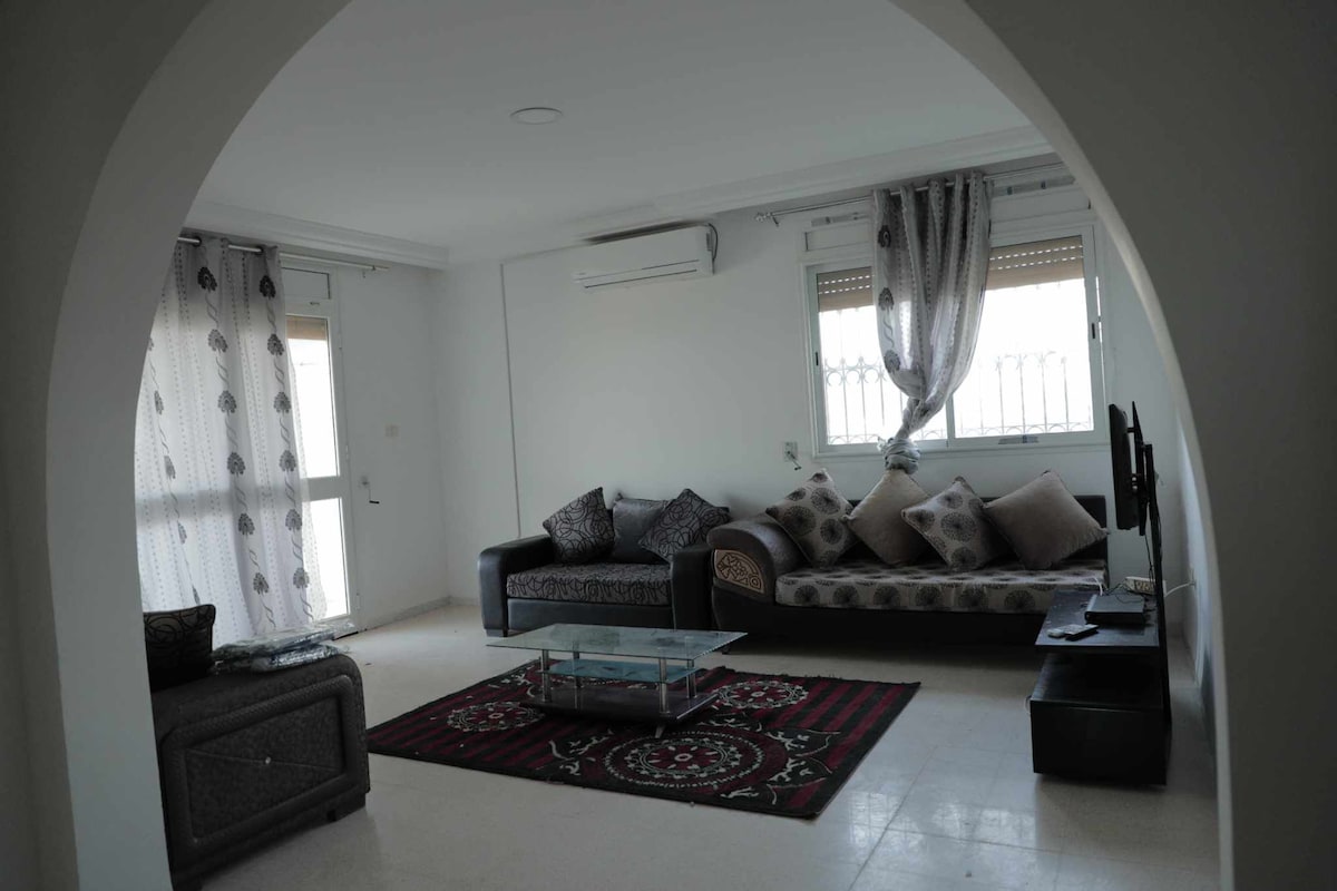 A spacious living area features a dark sofa adorned with patterned cushions, complemented by a glass coffee table at its center. A decorative rug lies underfoot, and light enters through large windows dressed with sheer curtains, enhancing the airy feel of the space.