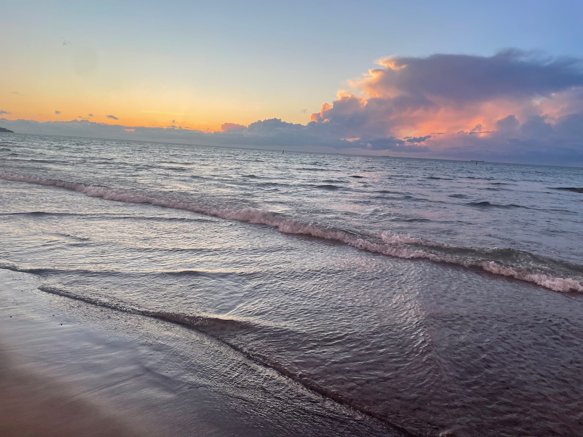 The image captures a serene beach scene at sunset, featuring gentle waves lapping against the shore. A pastel sky transitions from soft oranges to deeper blues, with clouds reflecting the warm light. The shoreline is visible in the foreground, creating a peaceful coastal ambiance.