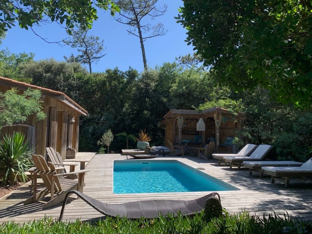 A wooden deck surrounds a clear swimming pool, complemented by sun loungers and outdoor seating. Lush greenery provides privacy, while a thatched-roof gazebo adds a tropical feel. A hammock rests nearby, enhancing the peaceful outdoor setting.