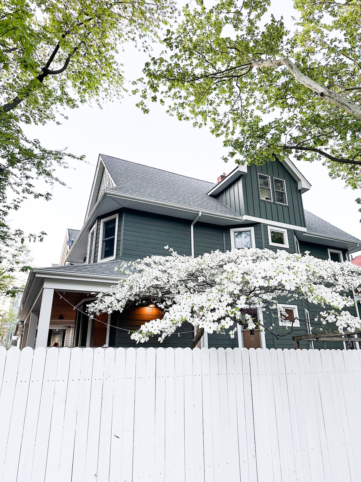 A charming house is framed by lush green trees and flowering branches, displaying a distinct architectural style. The exterior features a dark color palette and a gabled roof. A white picket fence enhances the inviting curb appeal, creating a welcoming entrance.
