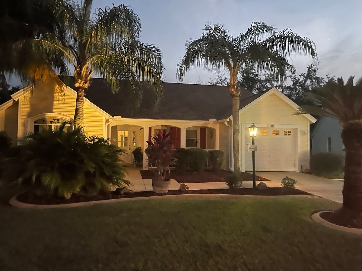 The exterior of a Monterey-style home is illuminated in soft evening light. Lush palm trees frame the entrance, leading to a covered porch. A well-maintained lawn features vibrant greenery, while a garage is visible to the right, contributing to the serene curb appeal.
