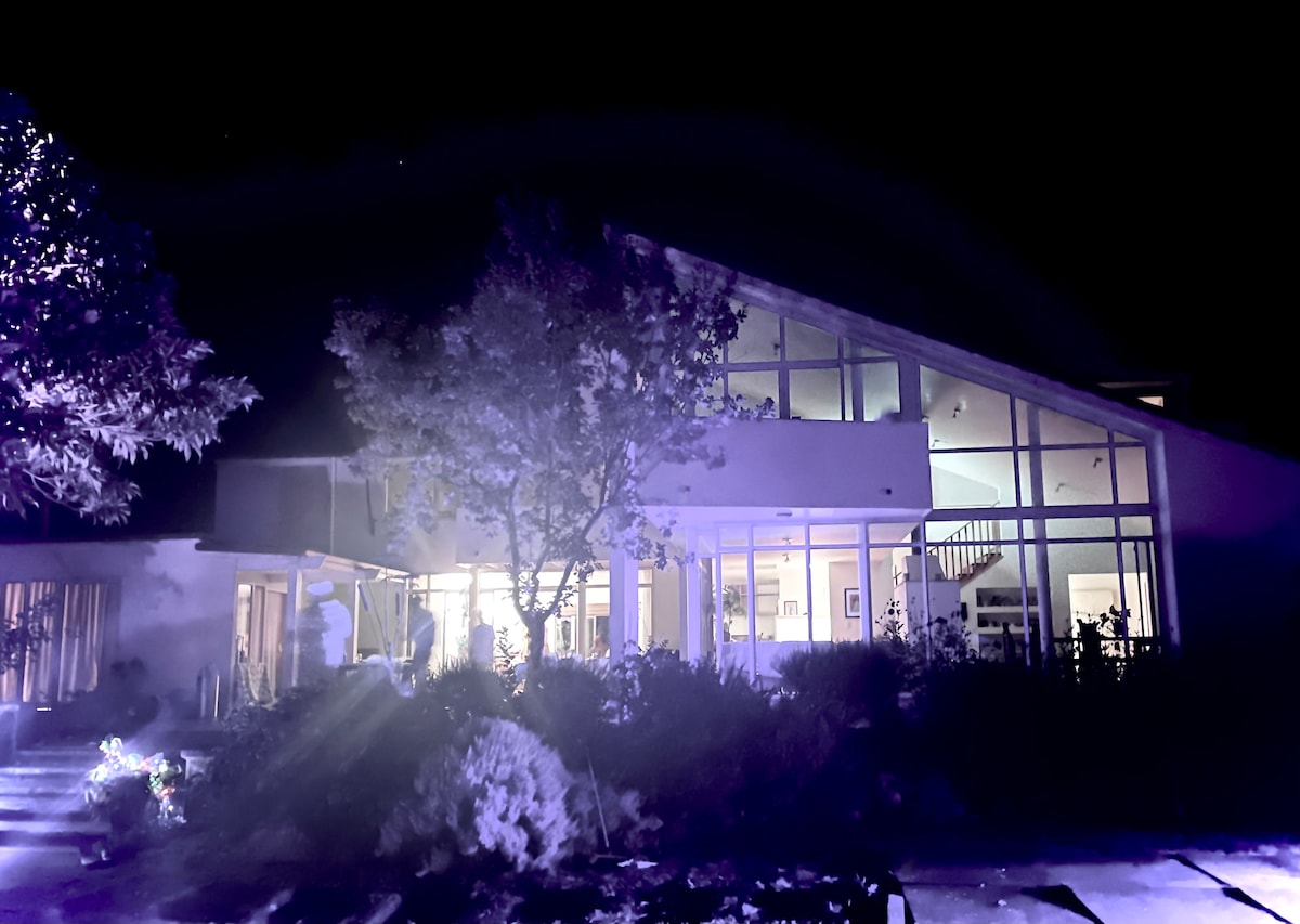 The exterior of the homestead is illuminated at night, showcasing its modern architecture and large glass windows. Surrounding greenery is visible, contributing to the serene atmosphere created by the soft lighting. A pathway leads towards the entrance, inviting exploration.