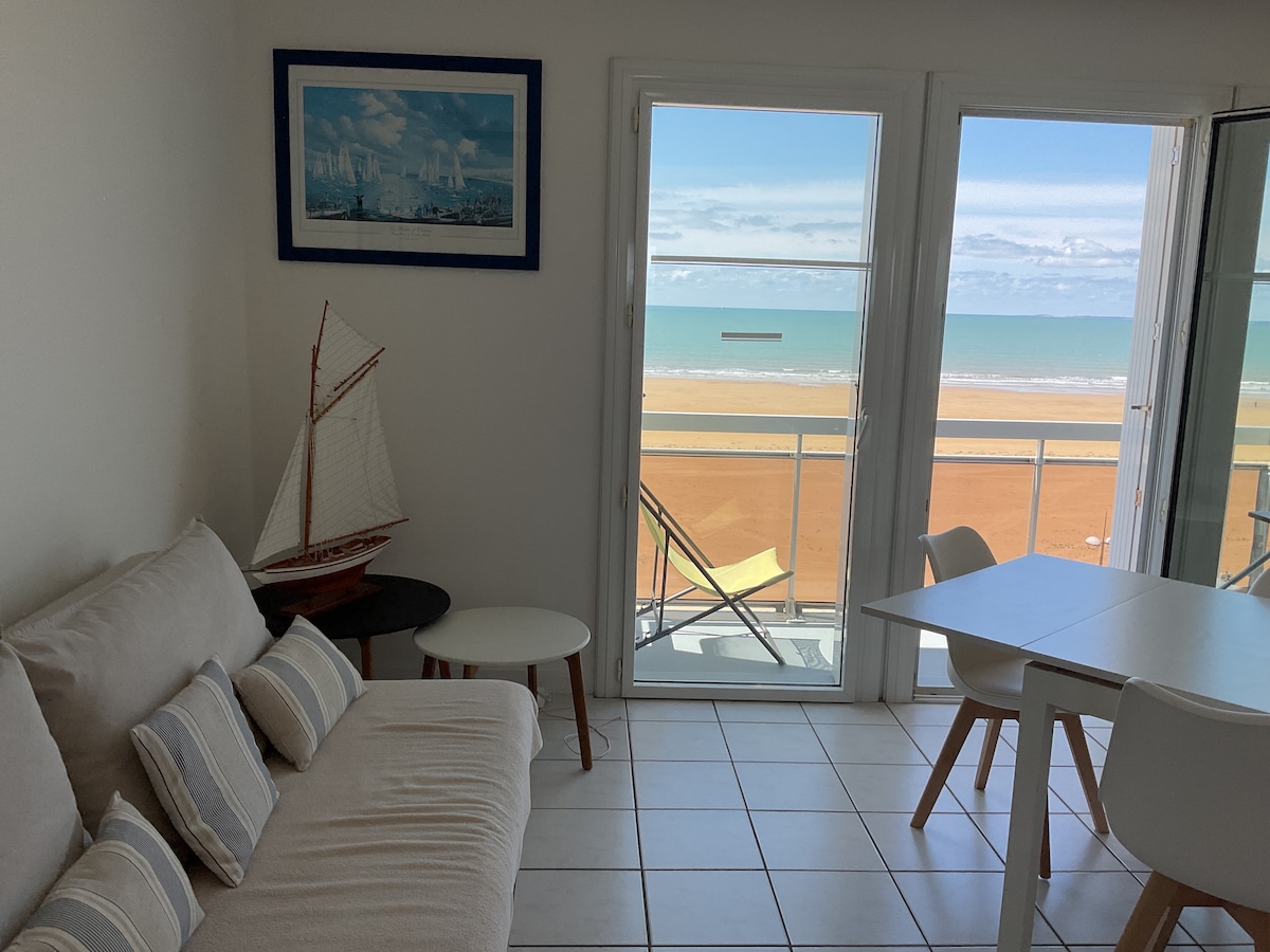 A bright living area features a neutral-toned sofa with decorative pillows, complemented by a small coffee table and dining table. Large glass doors open to a balcony, providing a clear view of the beach and sea, enhancing the space with natural light.