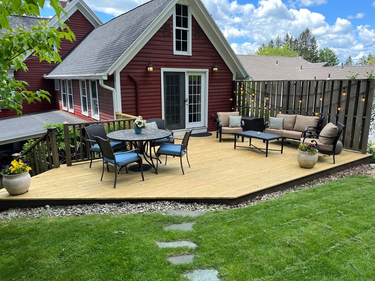 A spacious wooden deck is surrounded by green grass and features a seating area with a tan sofa, dark coffee table, and dining table set for six. Potted plants add greenery, while string lights provide soft illumination against the backdrop of the red home.