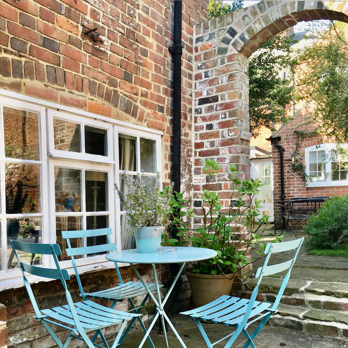 A quaint outdoor seating area is presented, featuring a round blue table accompanied by four matching chairs. The surrounding brickwork and archway add charm, while potted plants introduce greenery to the inviting space. Sunlight gently filters through the garden, enhancing the serene atmosphere.