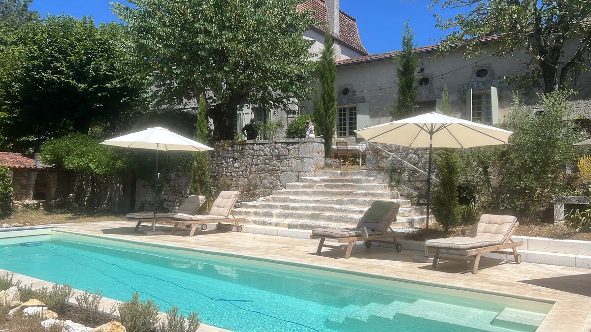 A serene swimming pool is framed by stone steps leading to a farmhouse. Sun loungers are positioned beside the pool, with two large umbrellas providing shade. Lush greenery surrounds the area, creating a private atmosphere, and the sky is clear and blue.