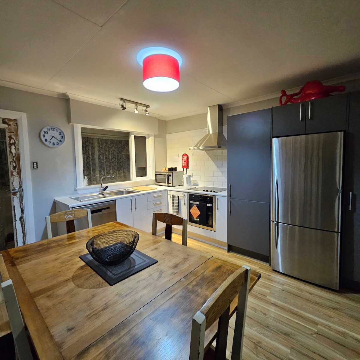 The kitchen features modern appliances, including a stainless steel refrigerator and oven, complemented by a sleek white backsplash. A wooden dining table with a central decorative bowl is positioned beneath a contemporary pendant light, offering a well-lit space for meal preparation and dining.