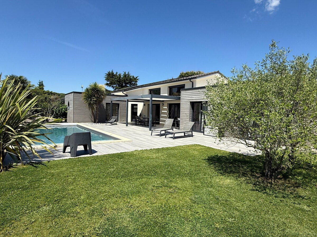 A modern, single-story house is surrounded by a verdant garden. The terrace, partially shaded by a pergola, features lounge chairs and a pool. Lush greenery and palm-like plants frame the scene, while clear blue skies provide a bright backdrop, showcasing the spacious outdoor area.
