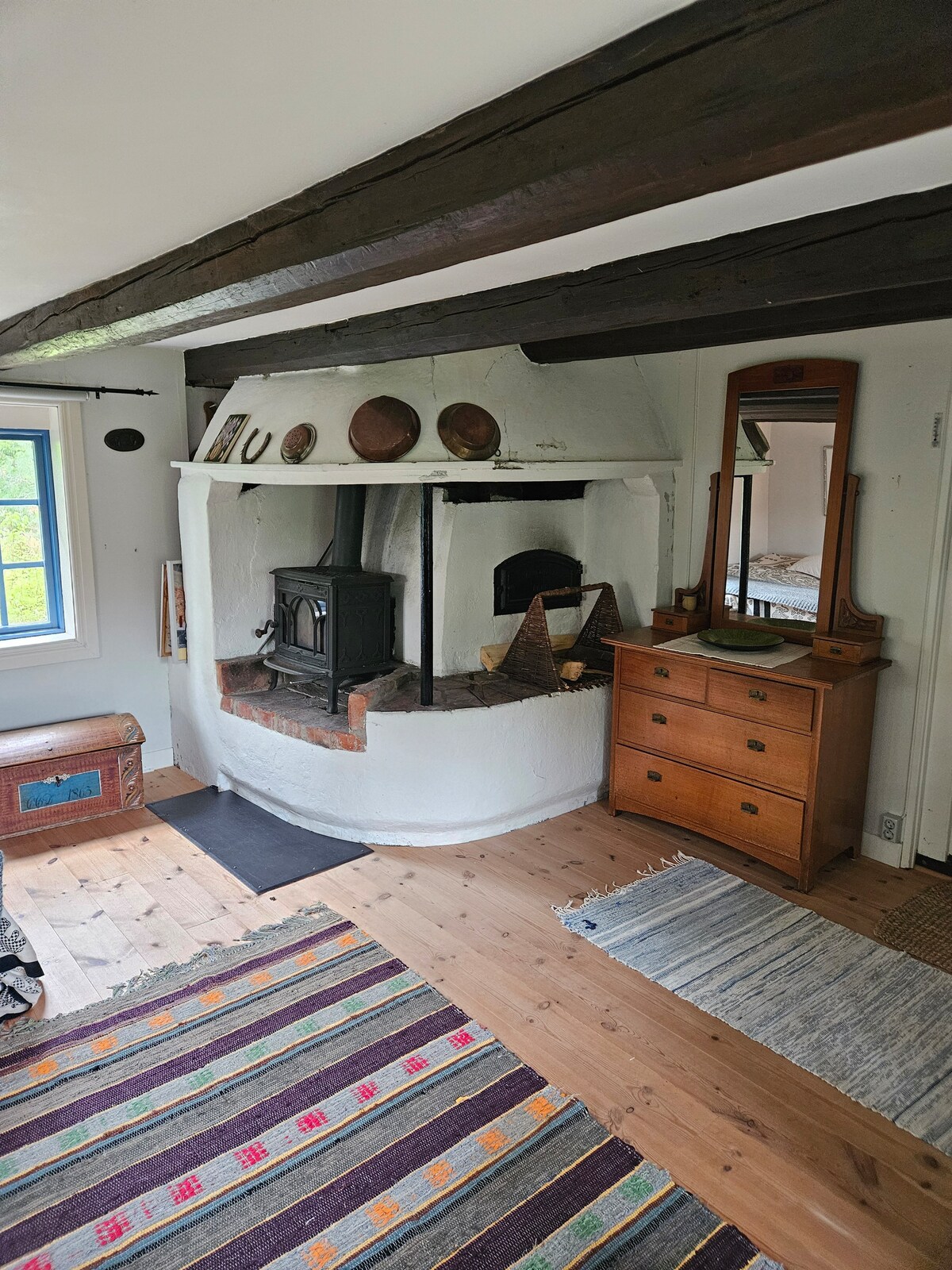 A cozy interior is highlighted by a traditional wood stove, set into a white-washed brick alcove. Natural light enters through the window, illuminating a wooden floor and a mix of area rugs. A wooden dresser and a small bench add to the warm atmosphere.
